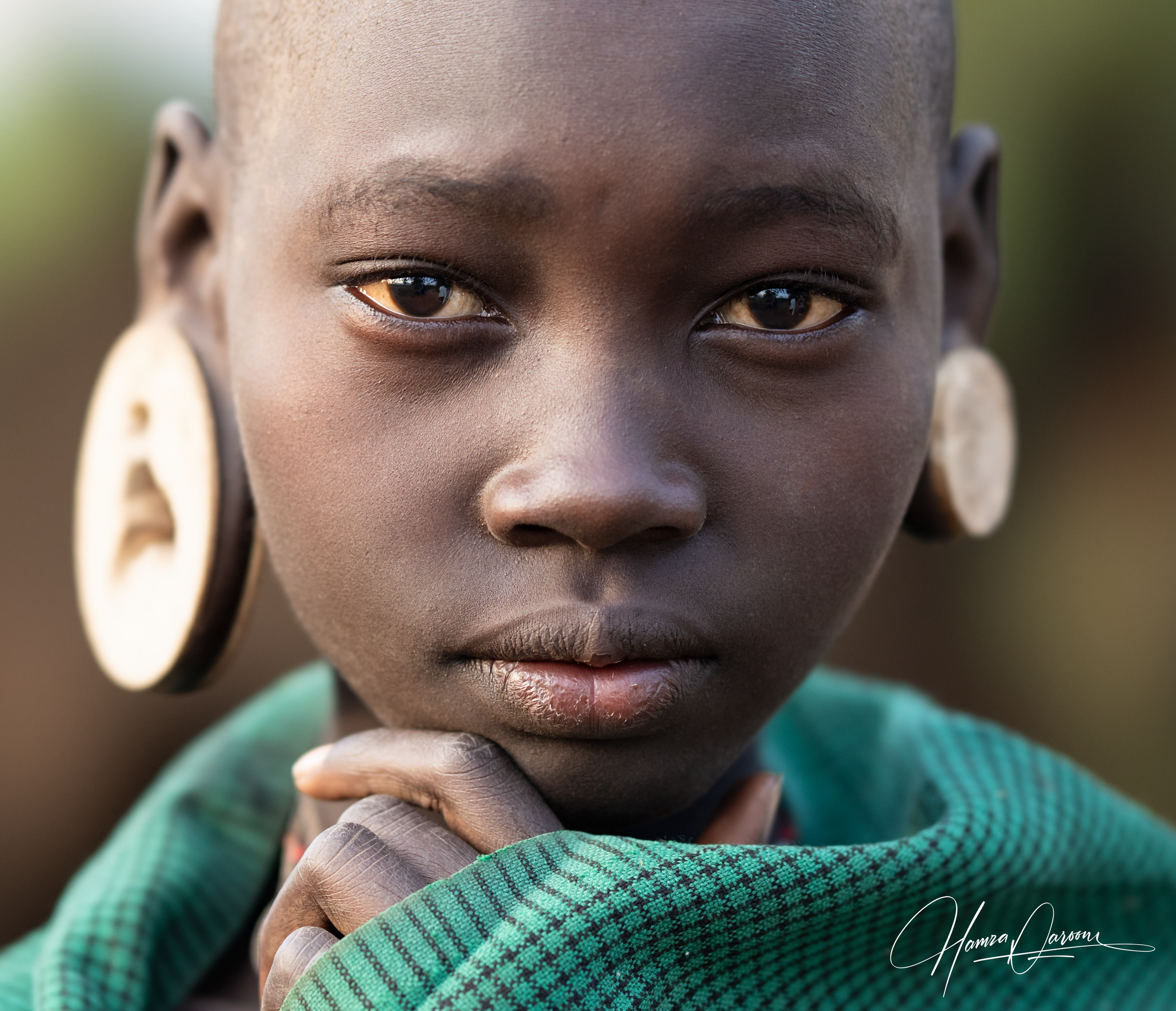Close-up of a young African girl with dark skin, large gold earrings, wearing a green and blue plaid shirt, looking directly at the camera with her hand touching her chin.