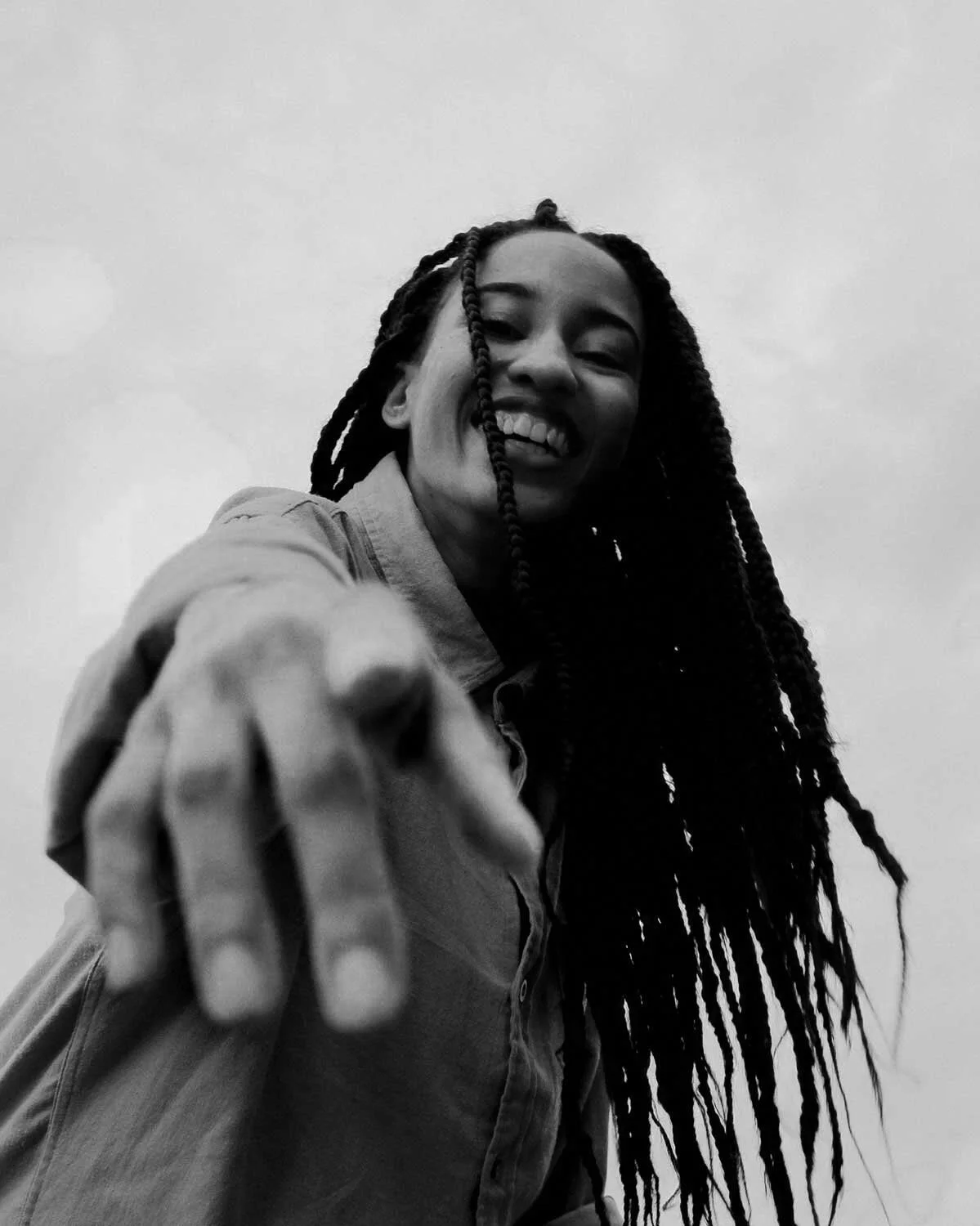 Black and white photo of a smiling woman with long braids reaching her chest, pointing towards the camera, with a plain sky background.