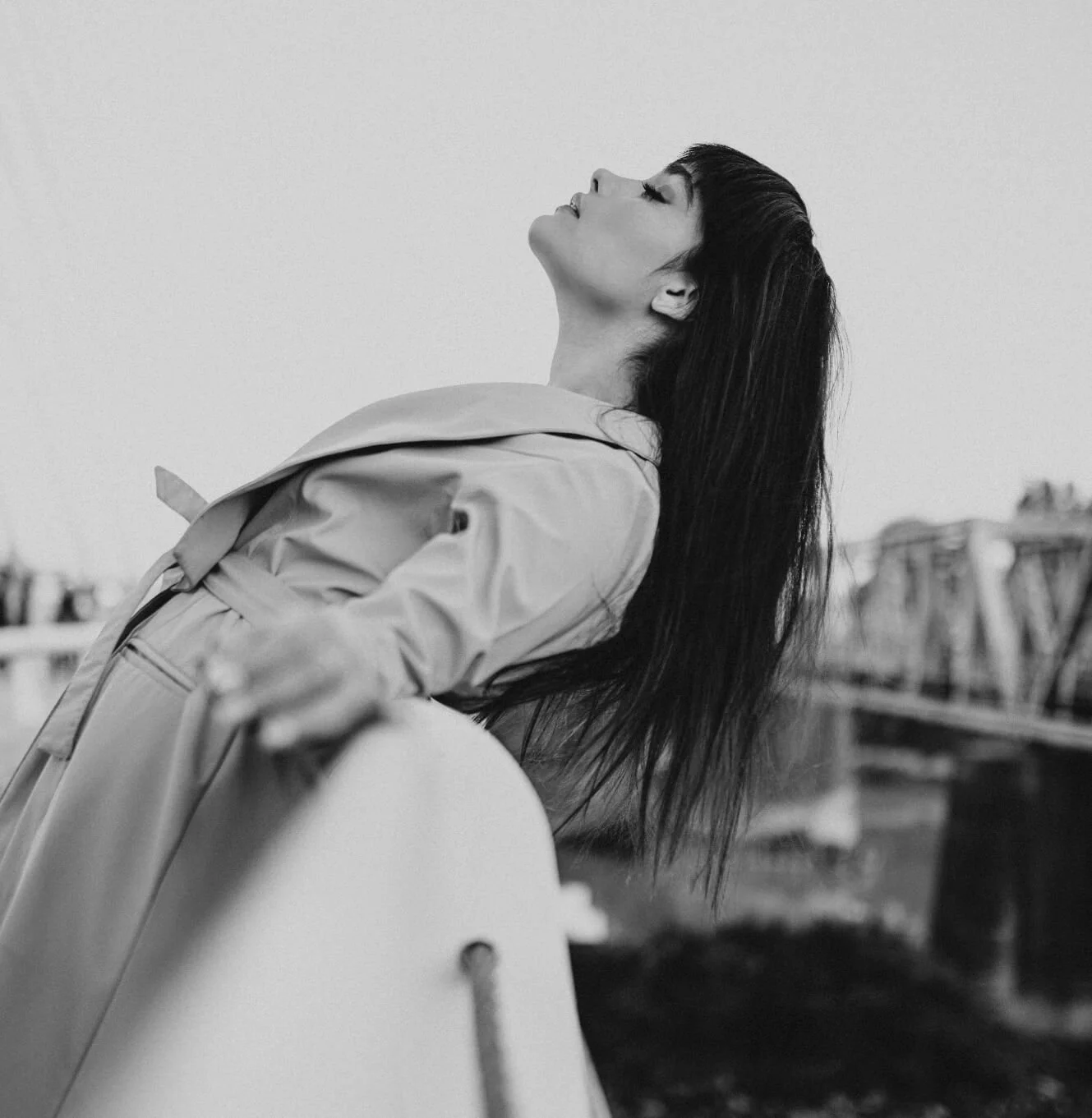 A woman with long dark hair and closed eyes leaning back with her face upward, wearing a light-colored trench coat, outdoors with a fence and buildings in the background.