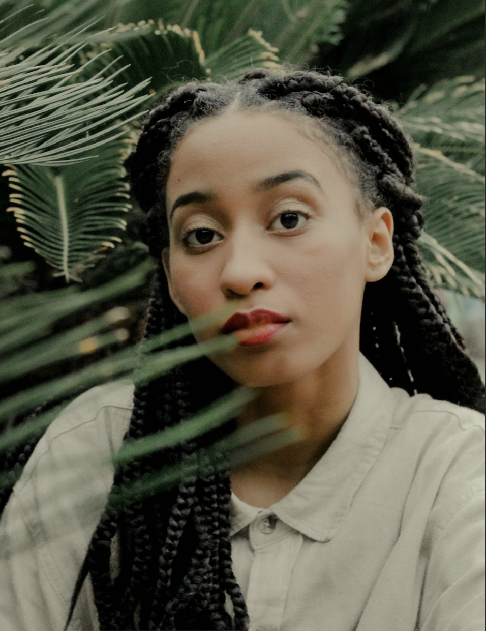 A young woman with braided hair and red lipstick looking at the camera surrounded by green tropical plants.