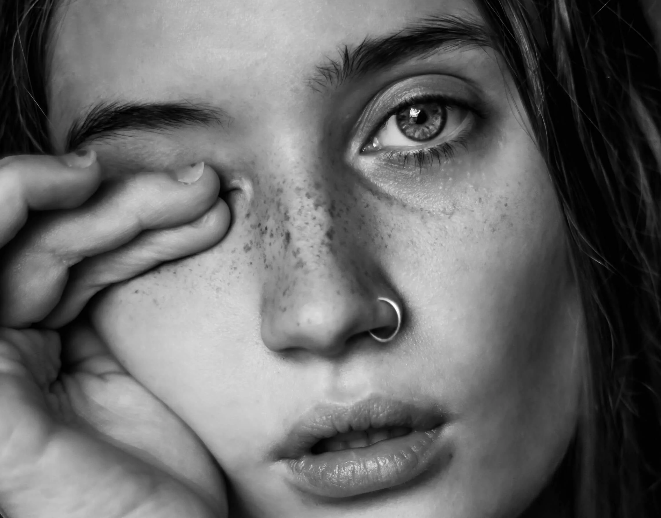 Close-up black and white photograph of a woman with a piercing in her nose, her chin resting on her hand near her lips, with her eye gazing into the distance.