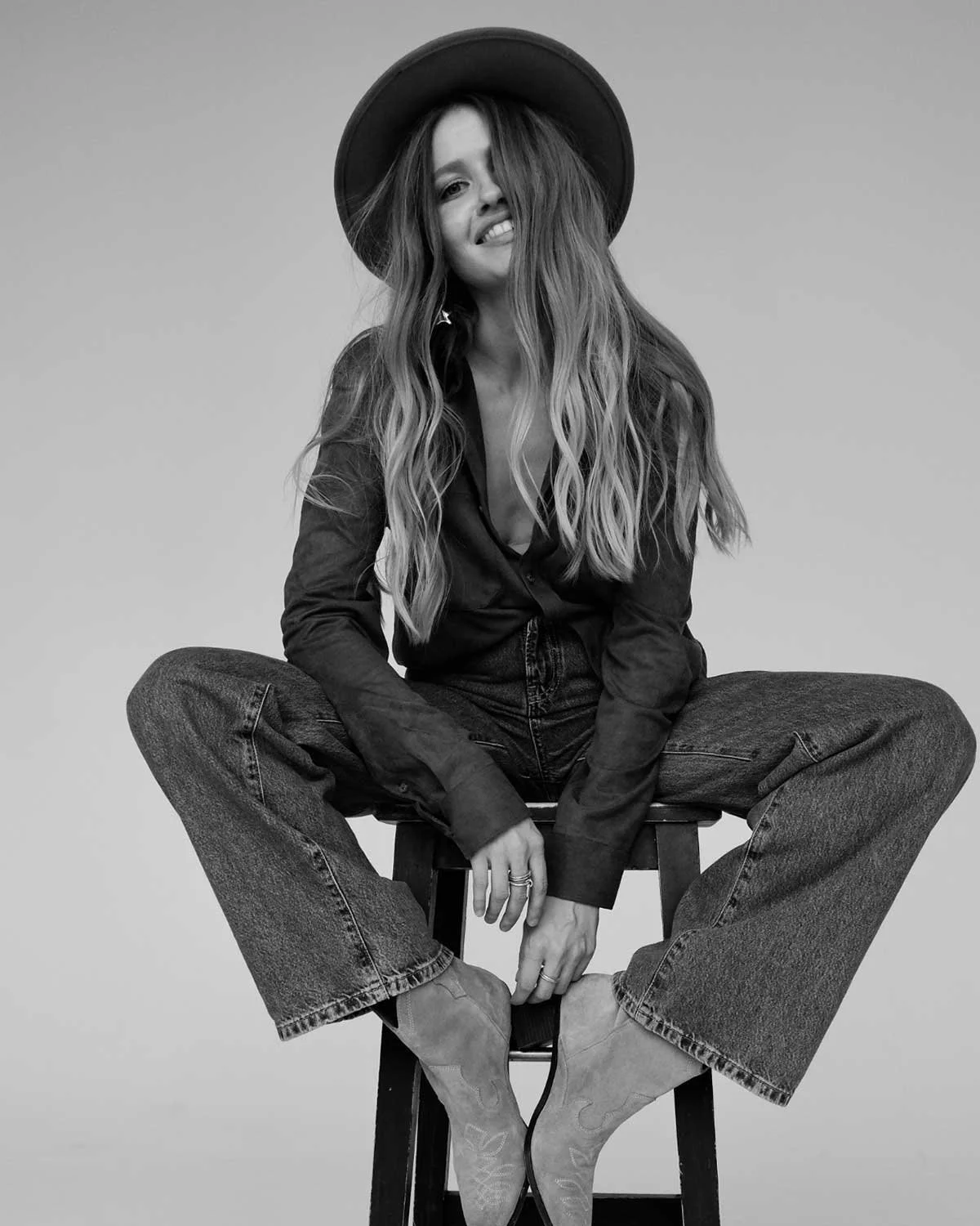 Black and white photo of a young woman sitting cross-legged on a stool. She has long, wavy hair, is wearing a wide-brimmed hat, a button-up shirt, jeans, and cowboy boots. She is smiling with one eye slightly closed and has a playful expression.