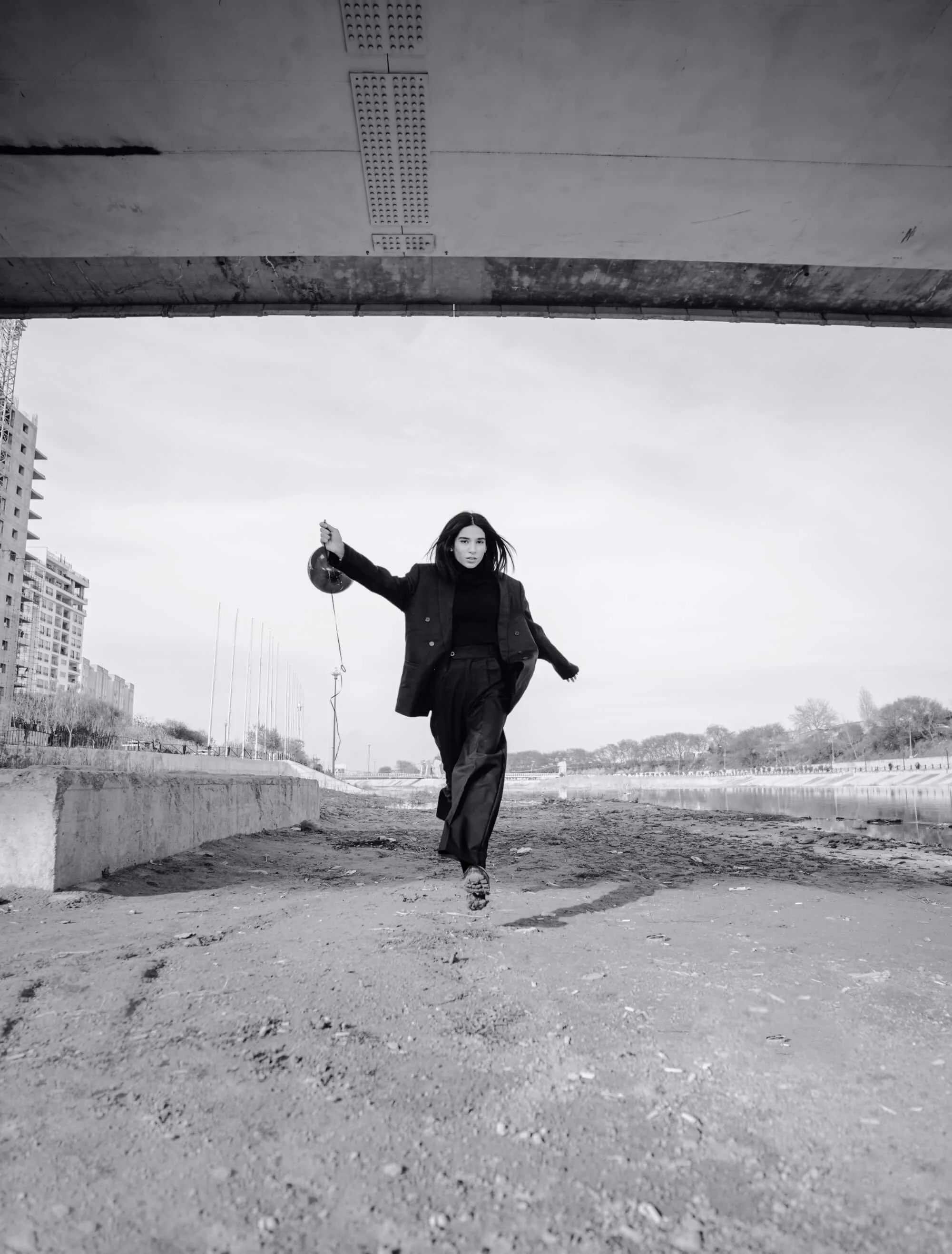 A woman in dark clothing, holding a balloon, walking on rough ground outdoors near a body of water and modern buildings, with a large concrete structure overhead.