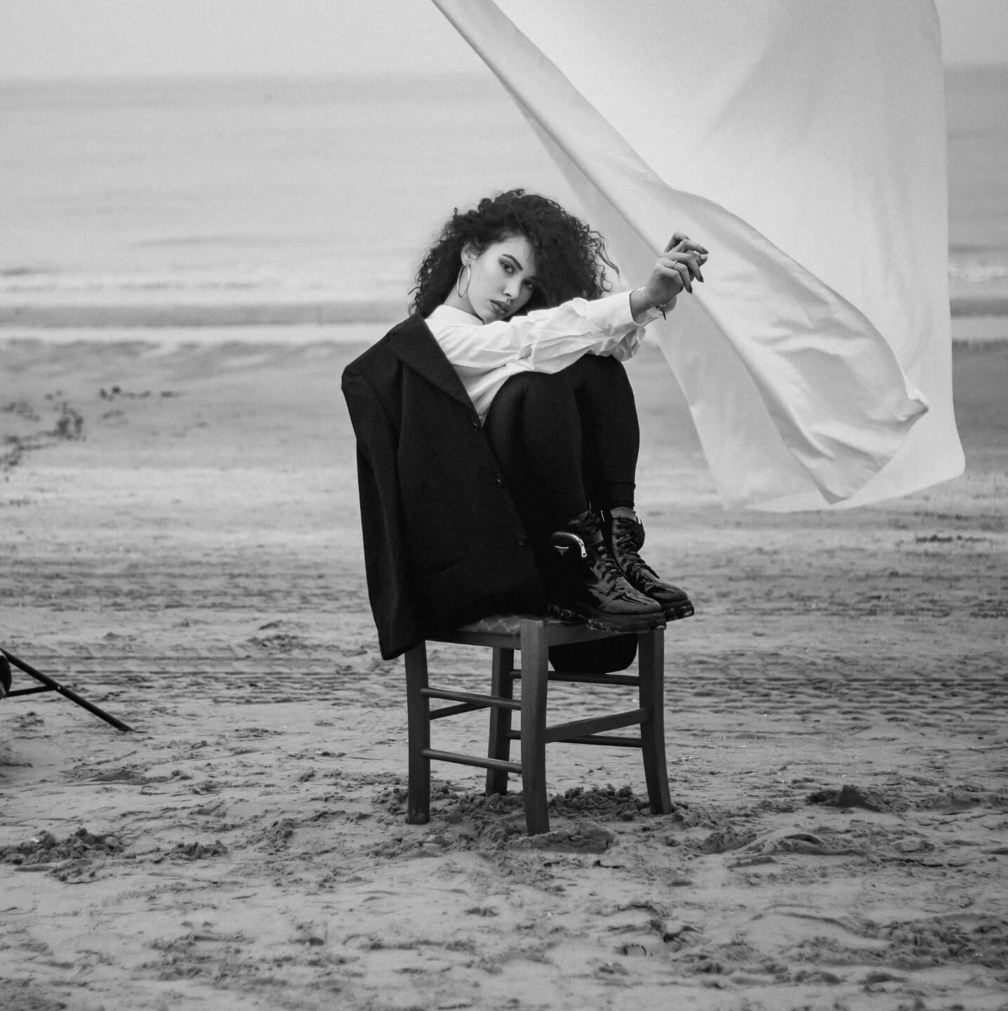 Woman with curly hair sitting on a chair on a beach, holding a large flag, dressed in a coat and boots, black and white photograph.