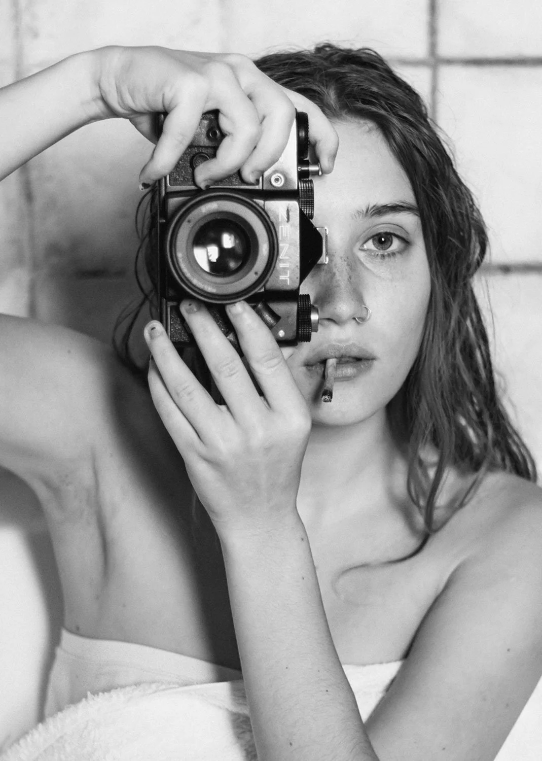 A young woman with wavy hair and a nose ring taking a black-and-white photograph with a vintage camera, holding a cigarette in her mouth, seated in front of a brick wall.