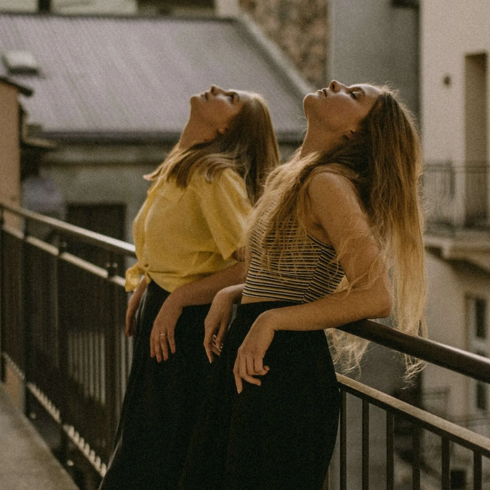 Two women sit on a balcony railing with their heads tilted back and eyes closed. They are illuminated by soft, warm lighting, with a brick wall in the background.