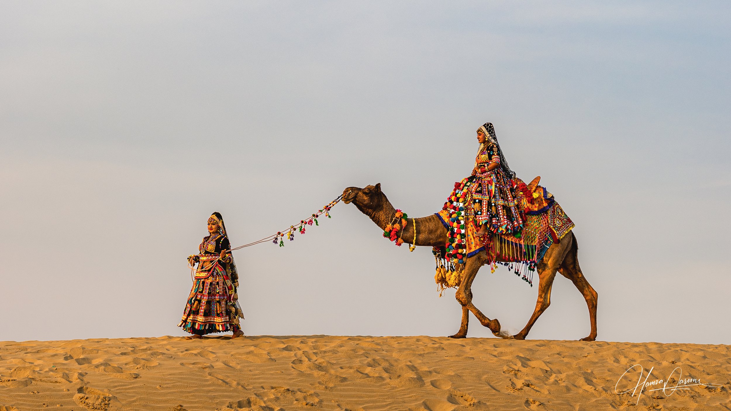 Two women dressed in colorful traditional Indian attire riding a decorated camel in a desert, with one woman leading the camel.