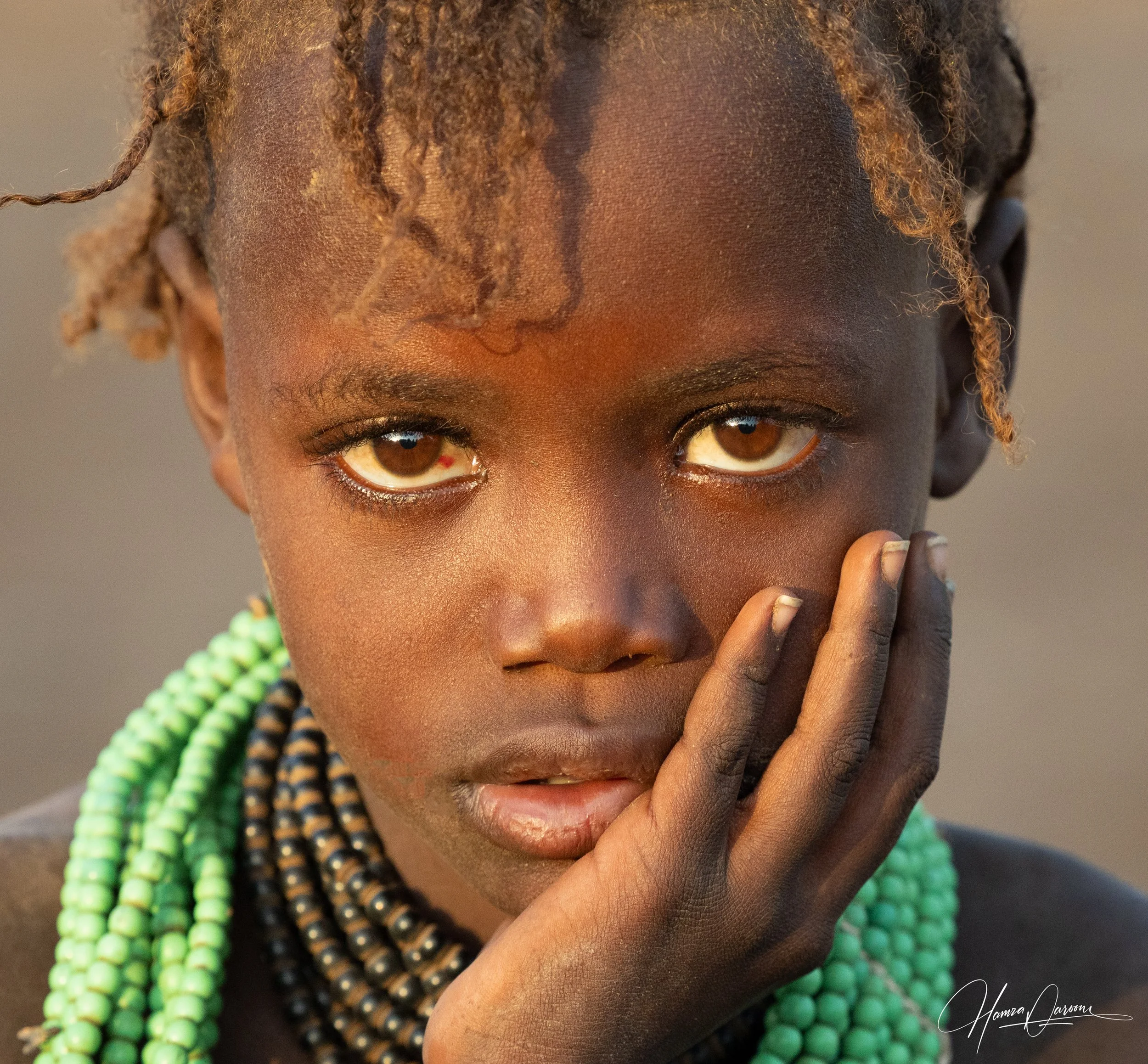 Close-up of a woman with dark skin and brown eyes, wearing multicolored beaded necklaces, touching her face with her hand.