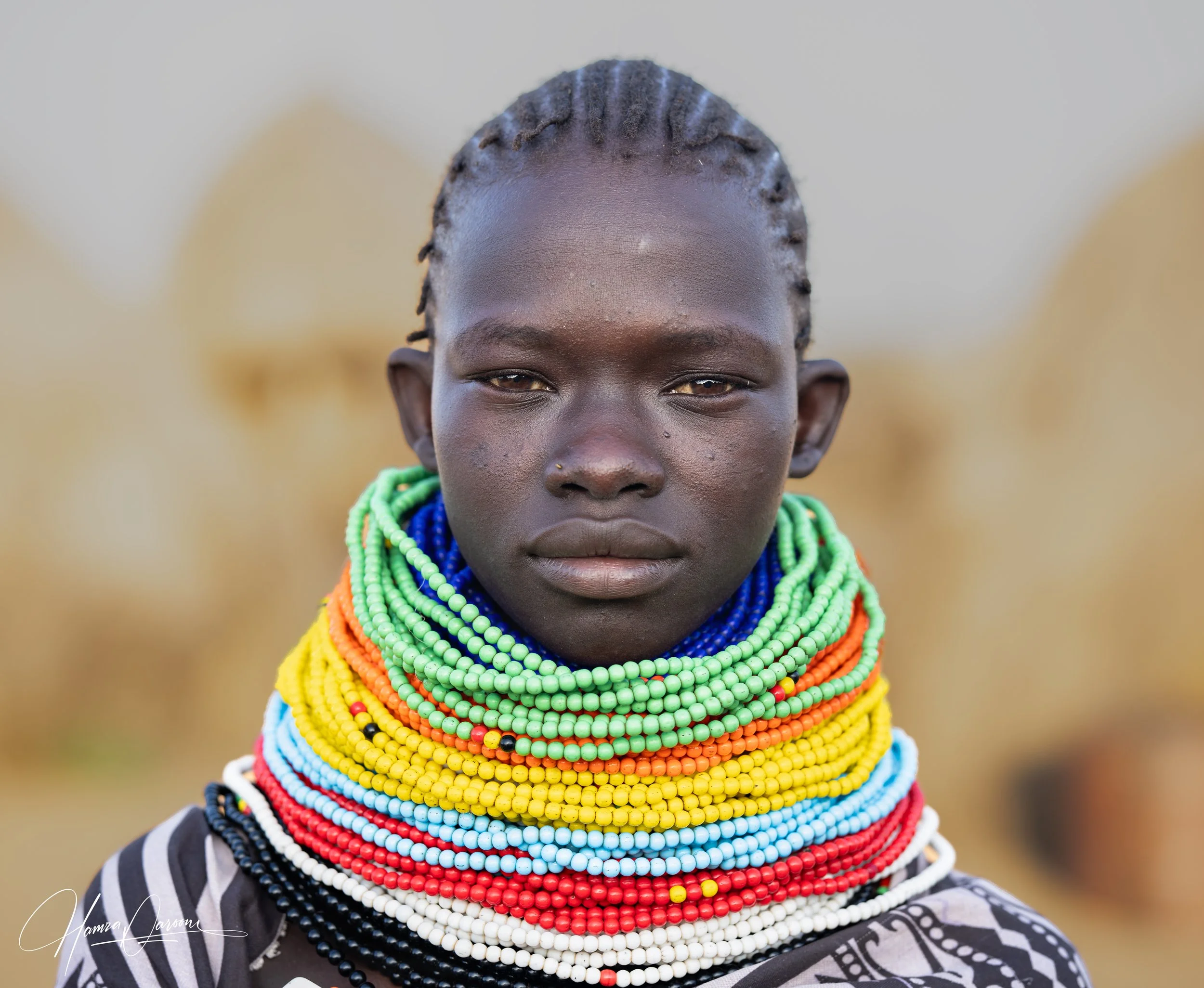 A young person with dark skin and braided hair wearing numerous colorful beaded necklaces and a black and white patterned shirt.