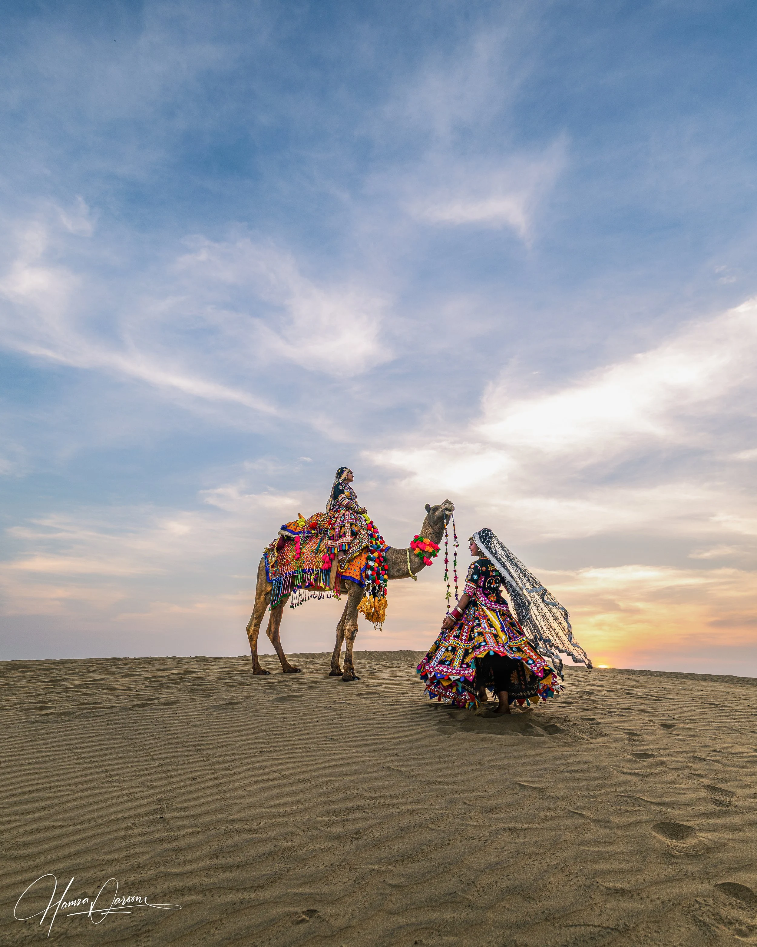 A woman in traditional colorful attire standing next to a decorated camel on desert sand during sunset.