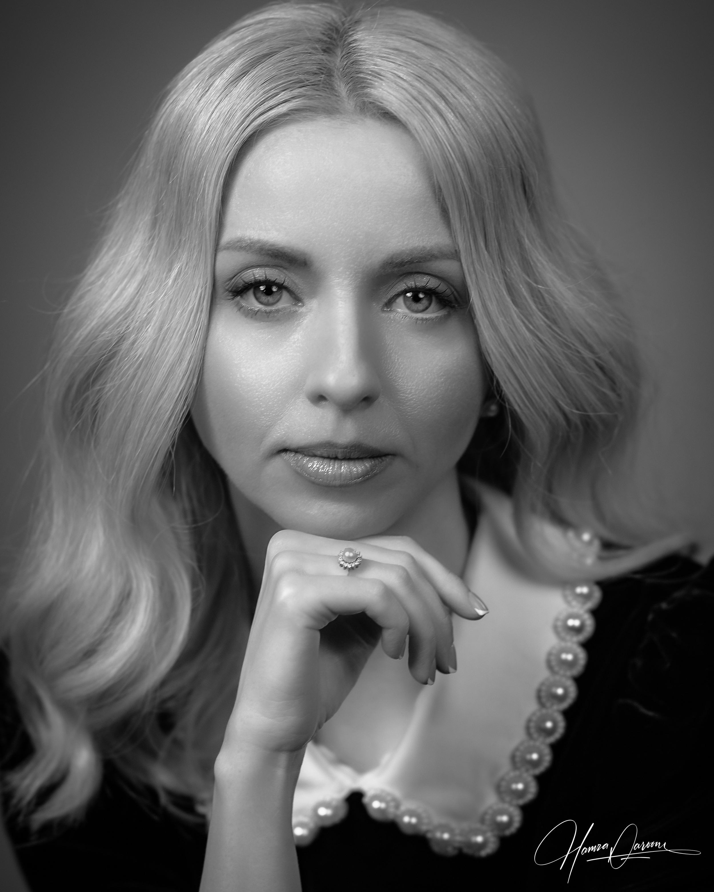 Black and white portrait of a woman with long, wavy hair, wearing a pearl bracelet and ring, looking directly at the camera with her hand near her chin.