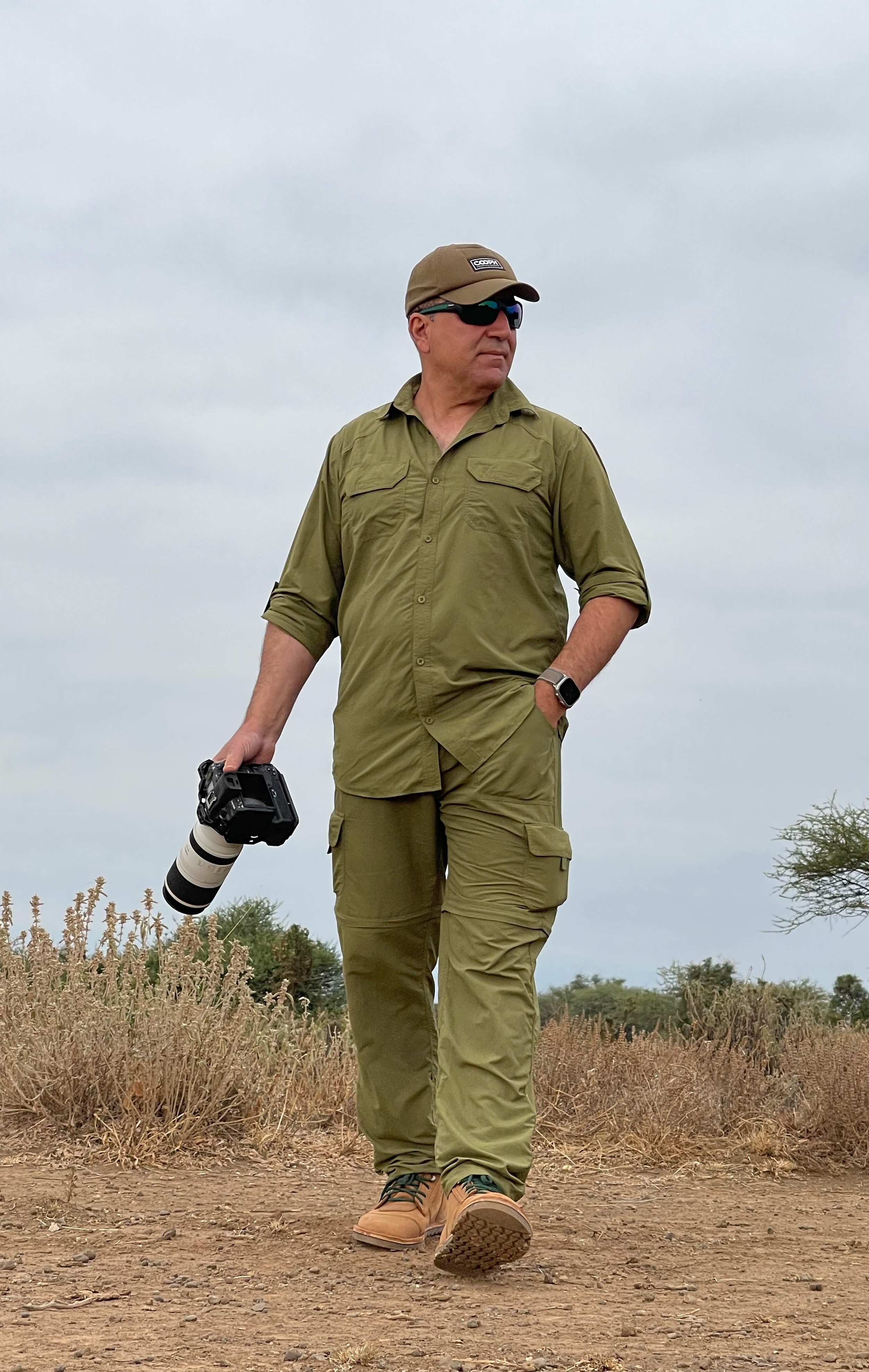 Man in tactical outdoor clothing walking on dirt trail, holding camera with telephoto lens, in a natural landscape under cloudy sky.