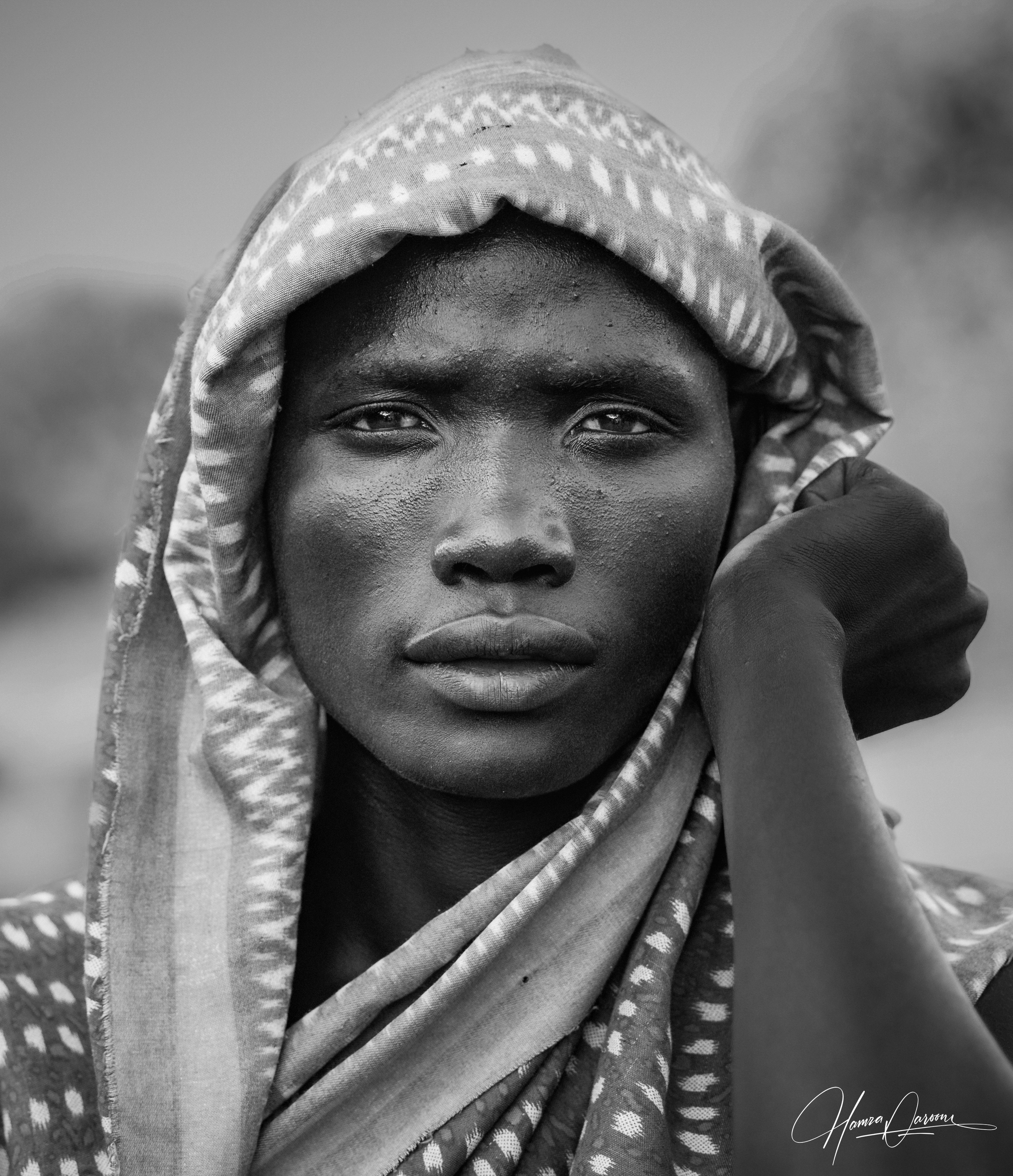 A close-up black and white portrait of a person with dark skin wearing a patterned head covering and traditional clothing, looking directly at the camera.