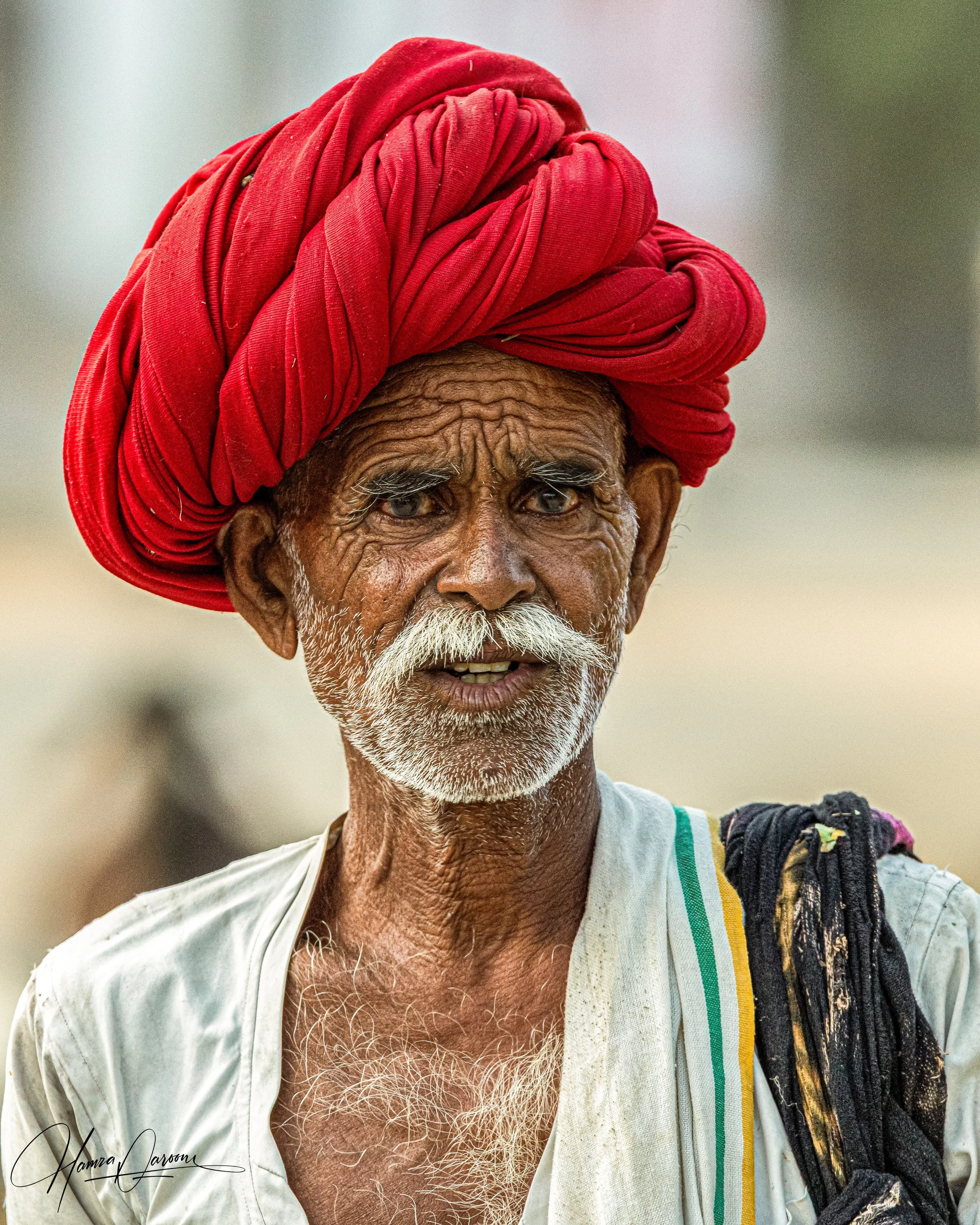 An elderly man with dark skin wearing a red turban, a white shirt with green and yellow stripes, and carrying a black bag, standing outdoors.