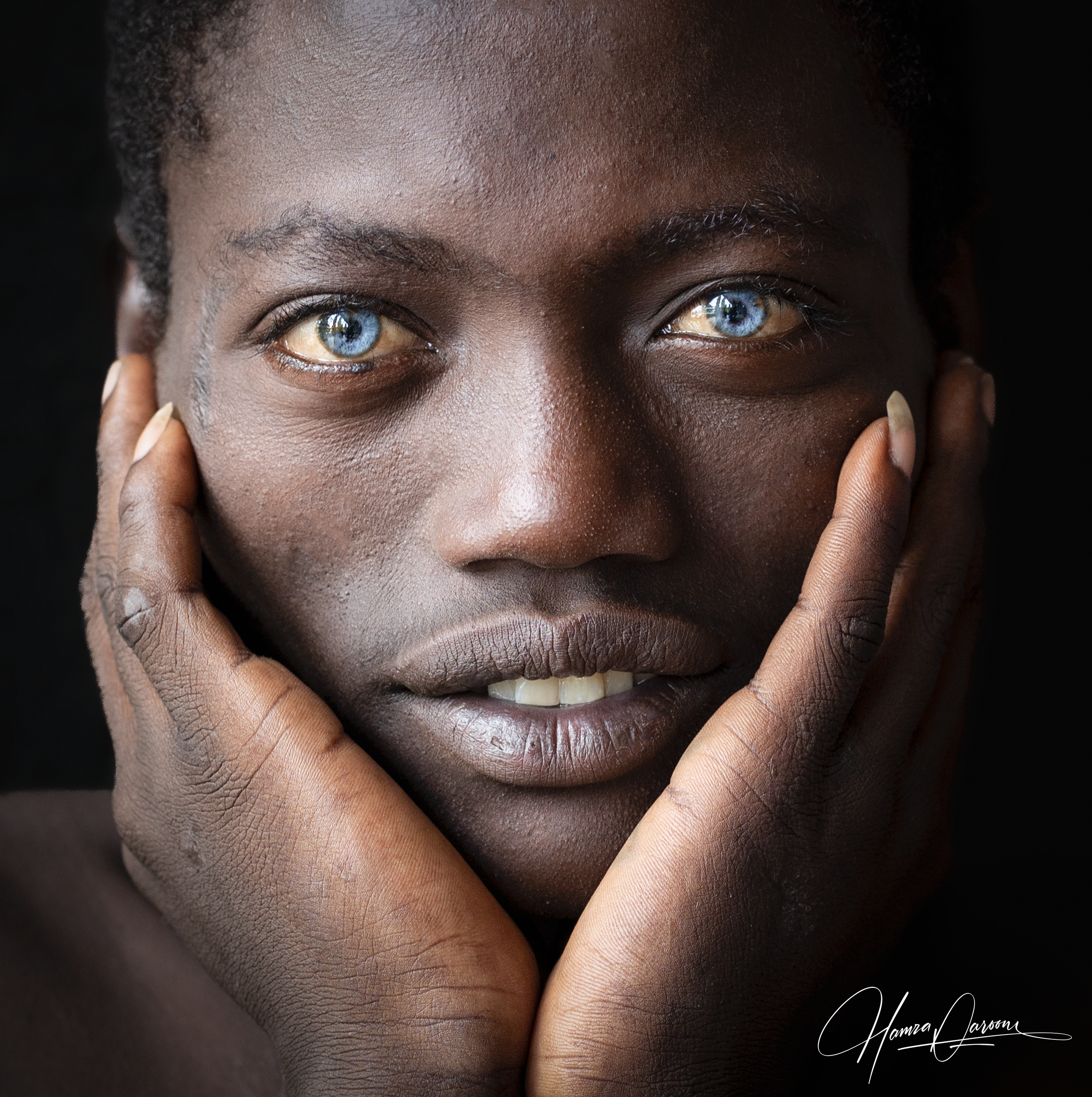 Close-up of a person with dark skin, blue eyes, and natural hair, smiling with fingers touching face, dark background.