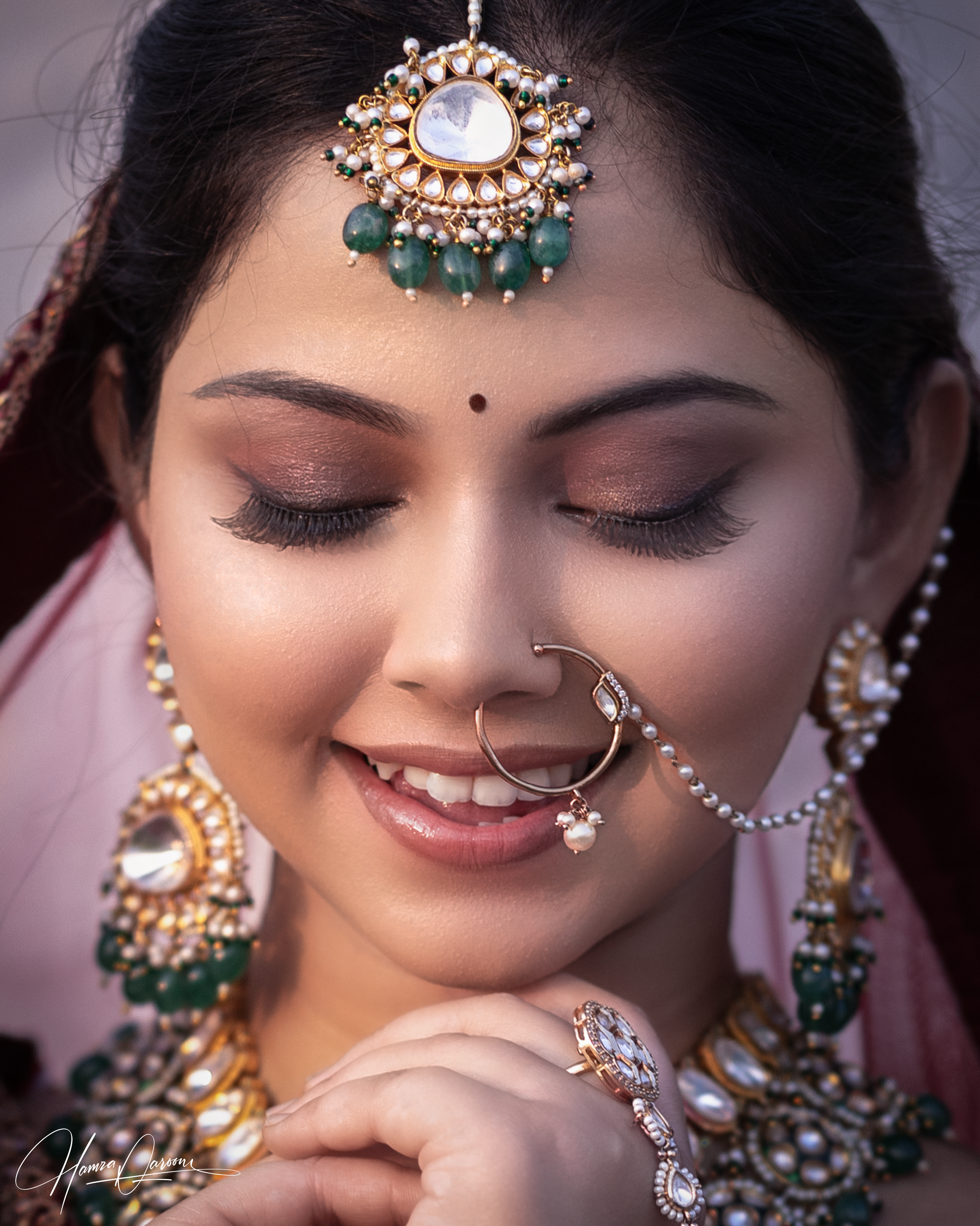 Close-up of a woman wearing traditional jewelry, including a large decorative headpiece with pearls and green beads, a nose ring, and earrings, all featuring white stones and gold accents. Her eyes are closed, and she is smiling slightly.