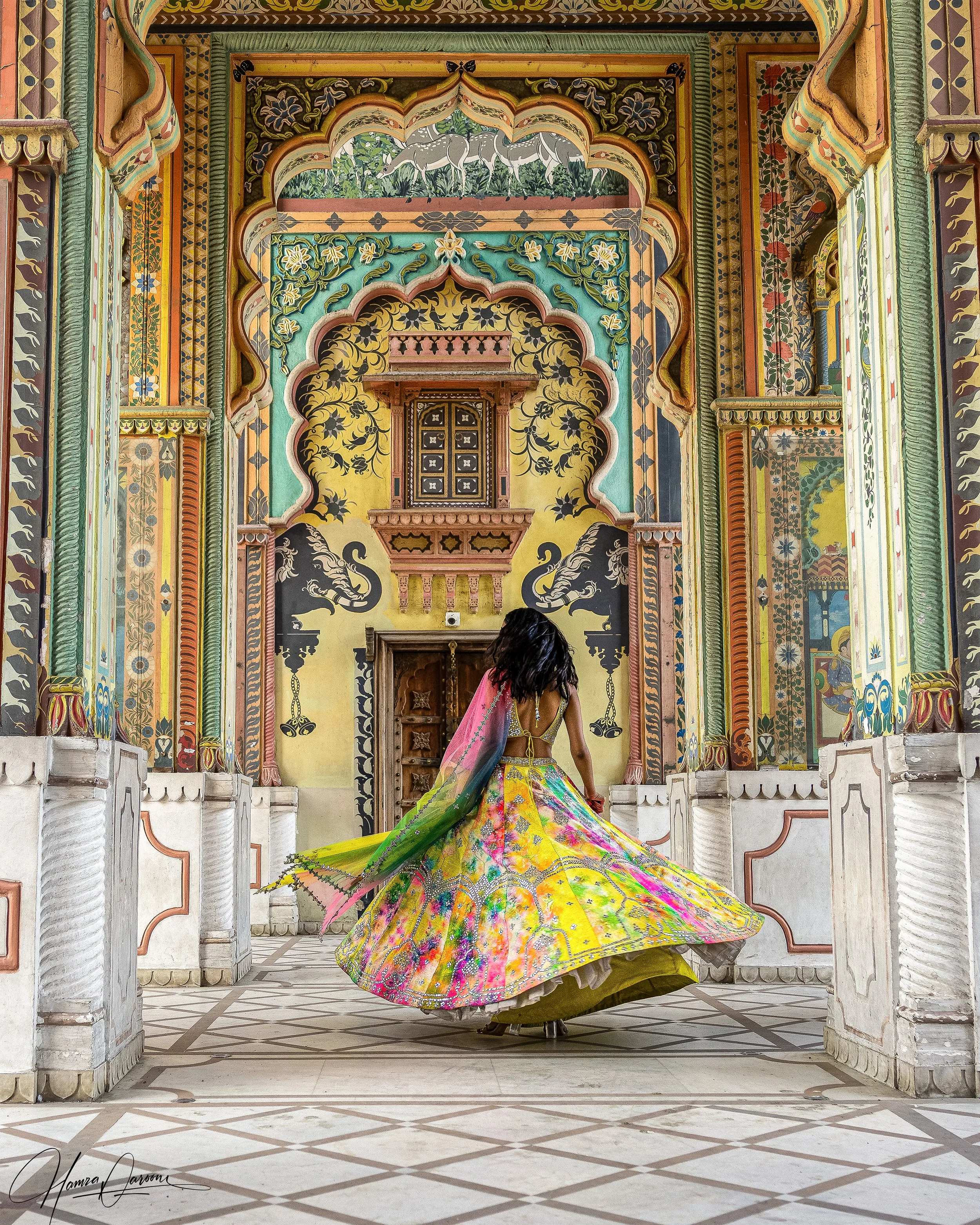 A woman in a colorful, flowing traditional dress is dancing inside a decorated palace with intricate wall paintings and arches. She is spinning, creating a wide, vibrant skirt.