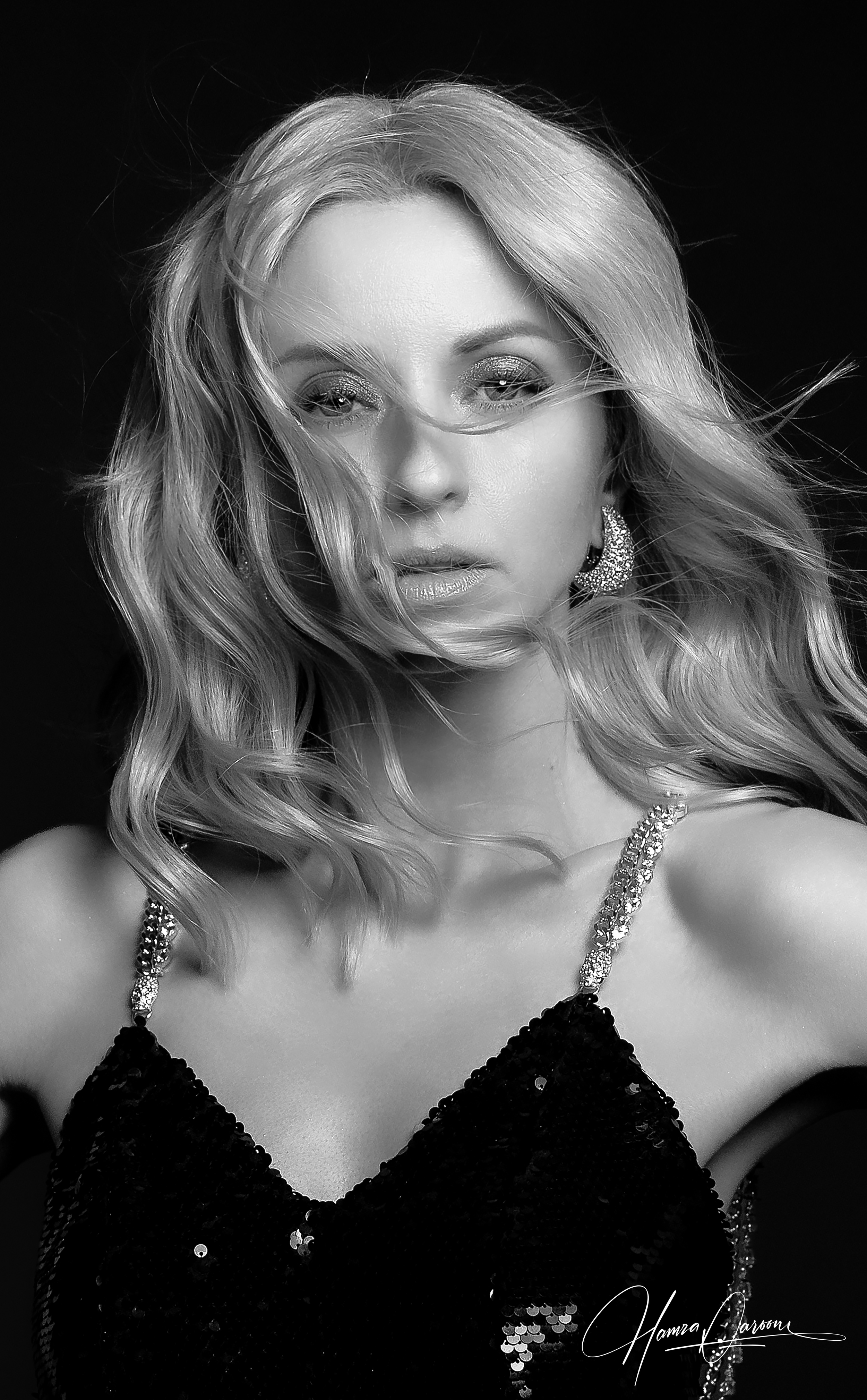 Black and white portrait of a woman with wavy hair, wearing sequined dress and jewelry, looking at the camera. Signature at the bottom right corner.