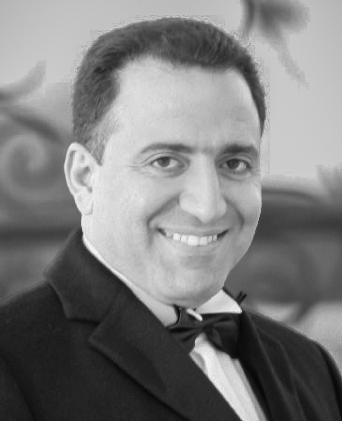 Black and white photo of a man in formal tuxedo and bow tie, smiling, with a blurred background.