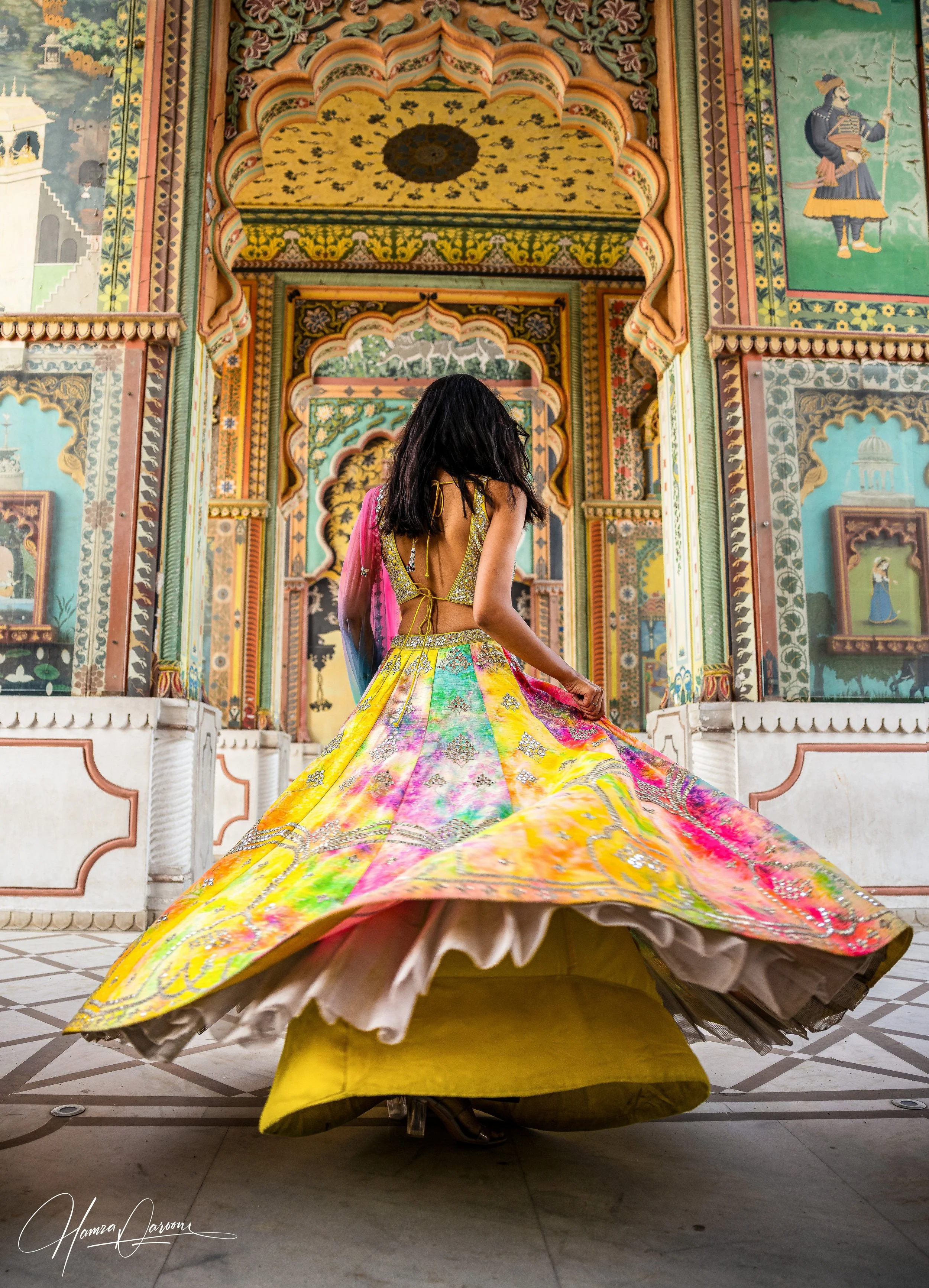 A woman in traditional Indian attire spinning a colorful lehenga inside an ornately decorated palace hall.