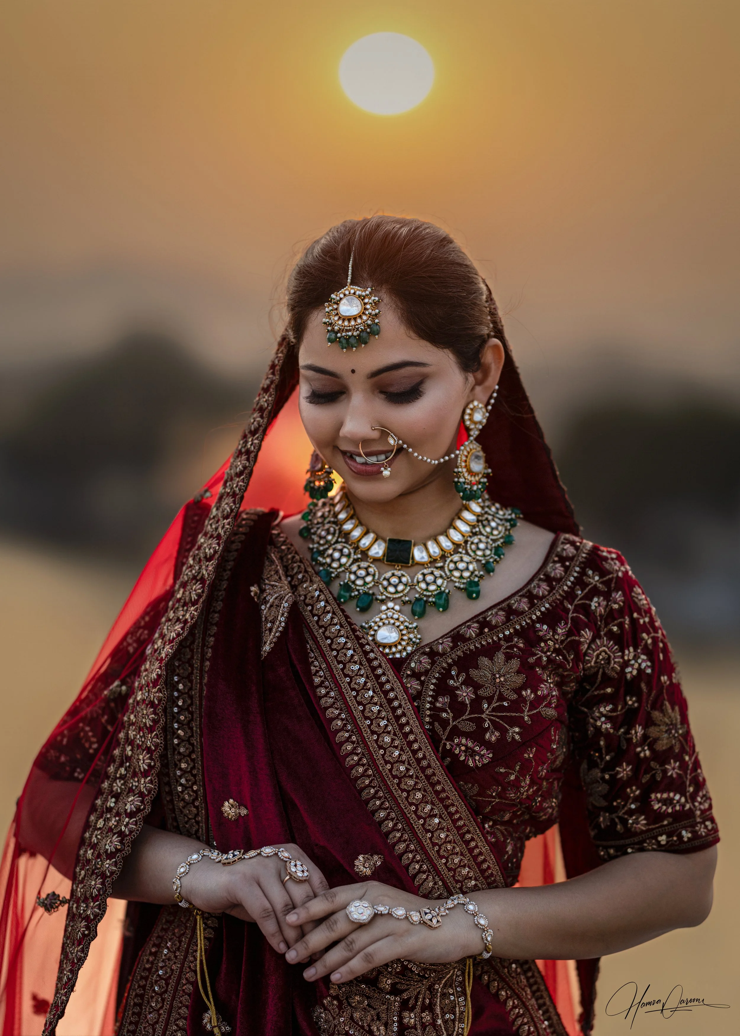 A woman dressed in traditional Indian attire, wearing elaborate jewelry including a maang tikka, earrings, necklace, bangles, nose ring, and rings, with a blurred sunset background.