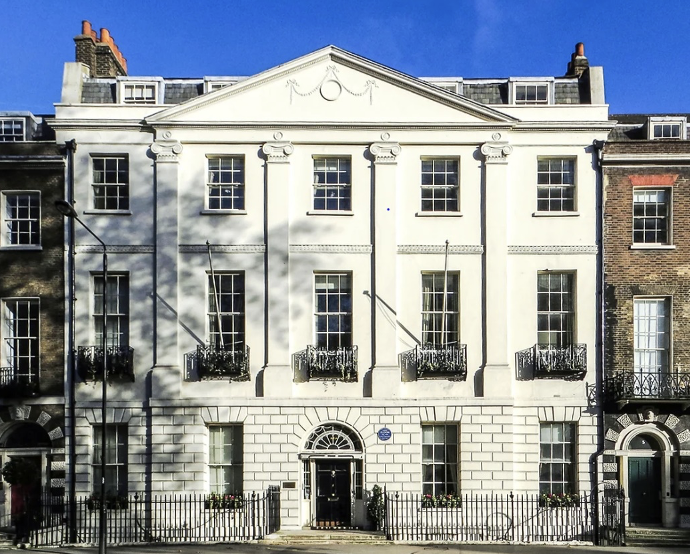 White neoclassical building with black iron balconies on upper floors, barred windows, and sculptural architectural details, surrounded by a fence, trees casting shadows, and neighboring buildings.