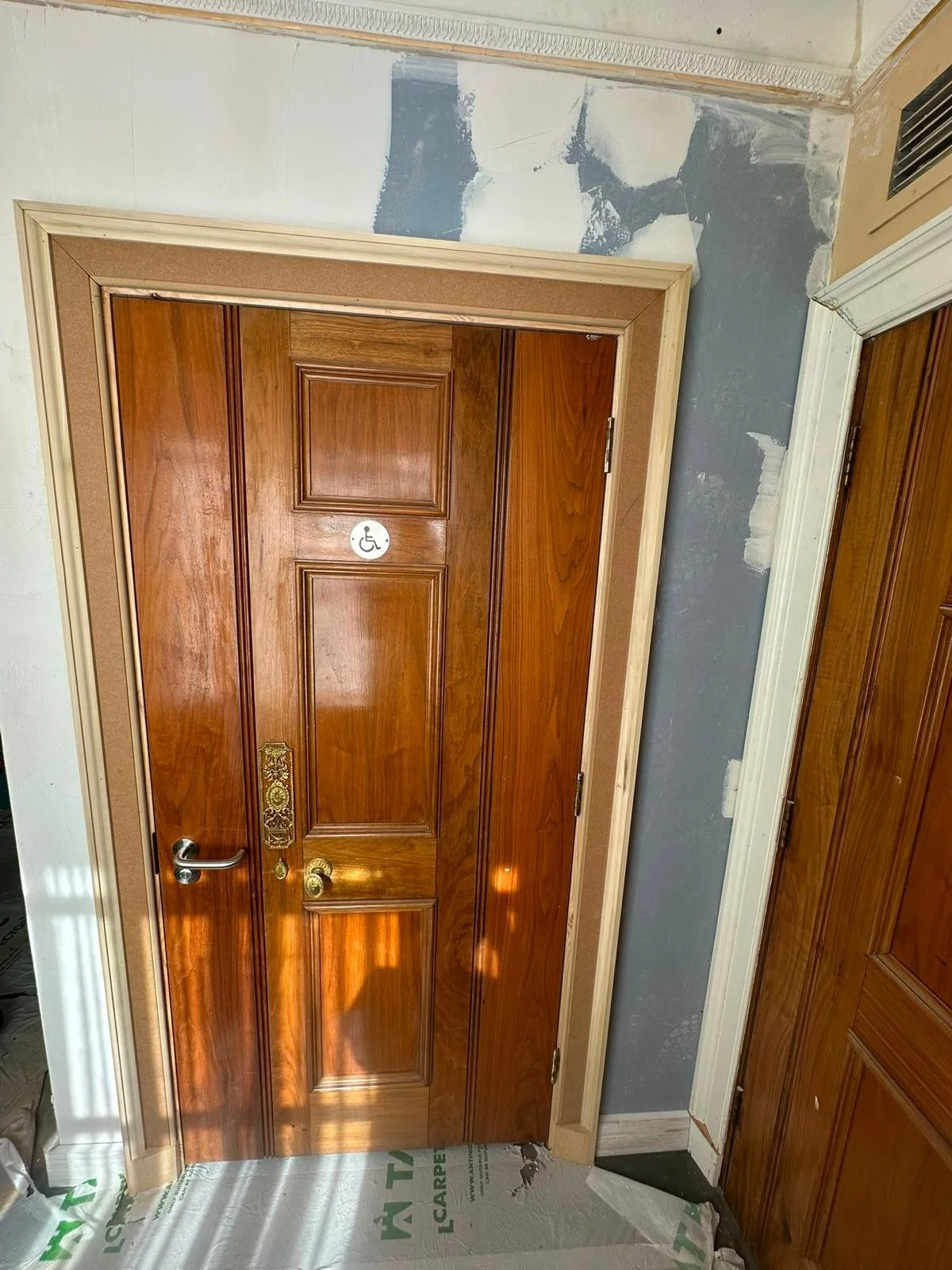 Wooden door with a small wheelchair accessibility sticker and a decorative brass lock, door frame surrounded by unfinished wall patchwork.