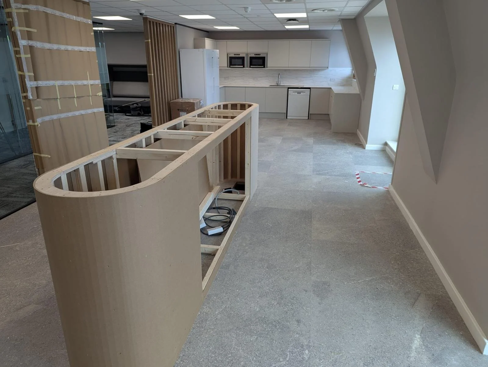 Construction in progress of a kitchen with unfinished cabinet framework, standing on a tiled floor, with a modern white kitchen area in the background and construction tape on the floor.
