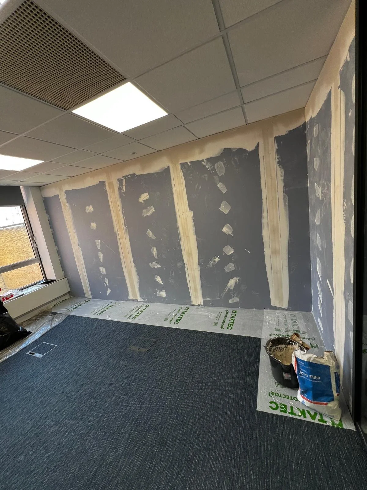 An unfinished interior room undergoing renovation, with walls partially covered with drywall and insulation, and a blue carpeted floor with construction materials on it.