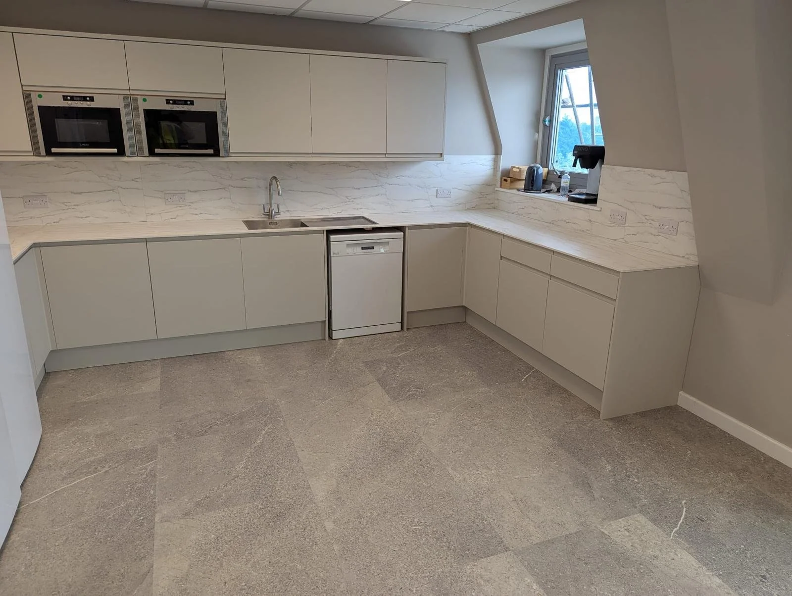 Modern kitchen with beige cabinets, marble countertops, a sink with a faucet, a built-in dishwasher, and a window with a view outside. Appliances include a microwave, oven, kettle, and coffee machine.
