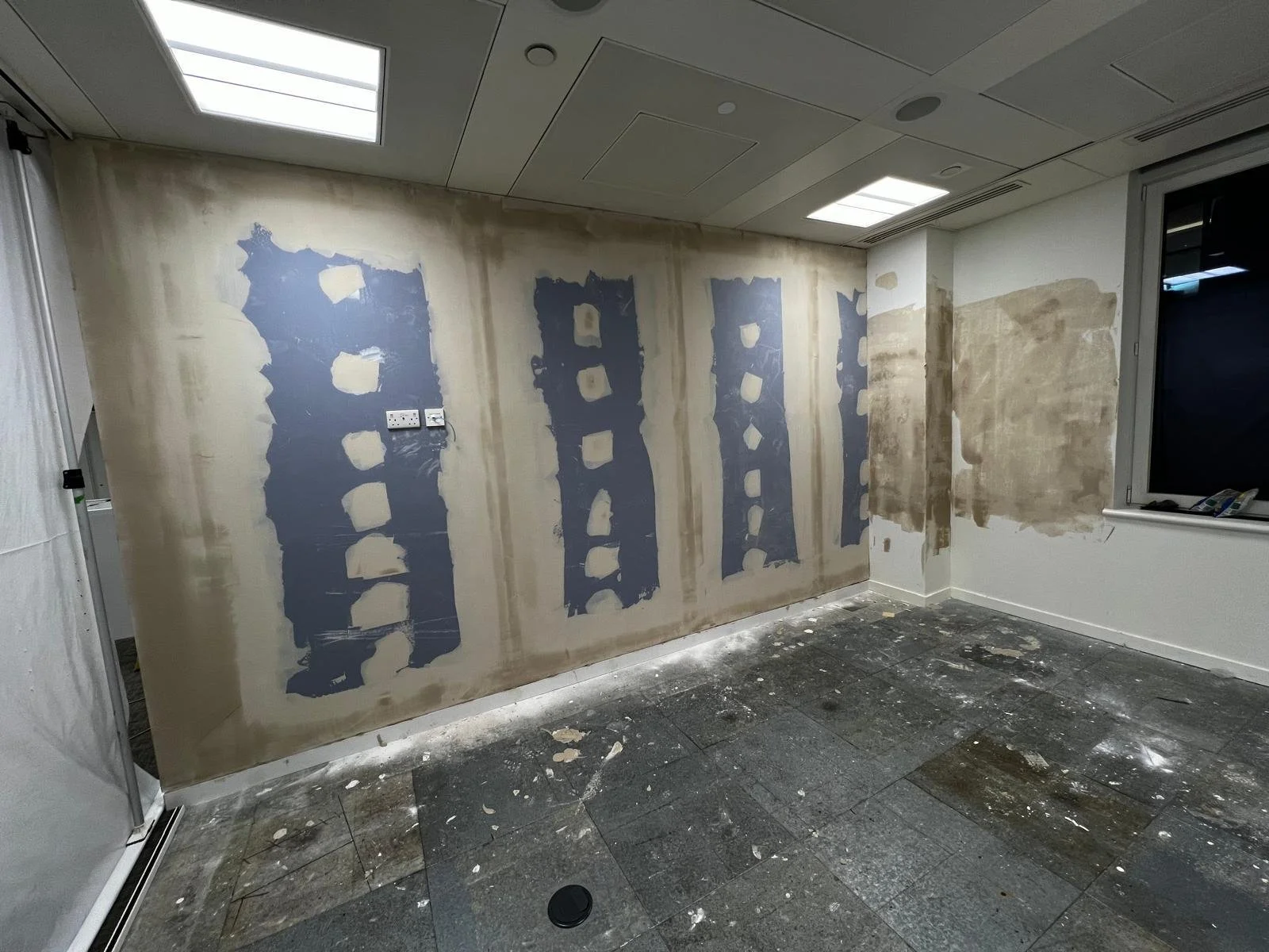 A room under renovation with partially painted walls and construction debris on the floor, featuring ceiling lights and a window.