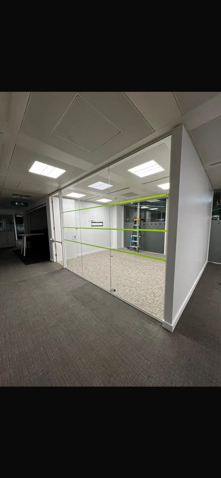 Empty office room with glass walls, green tape lines, ladder, and ceiling lights.