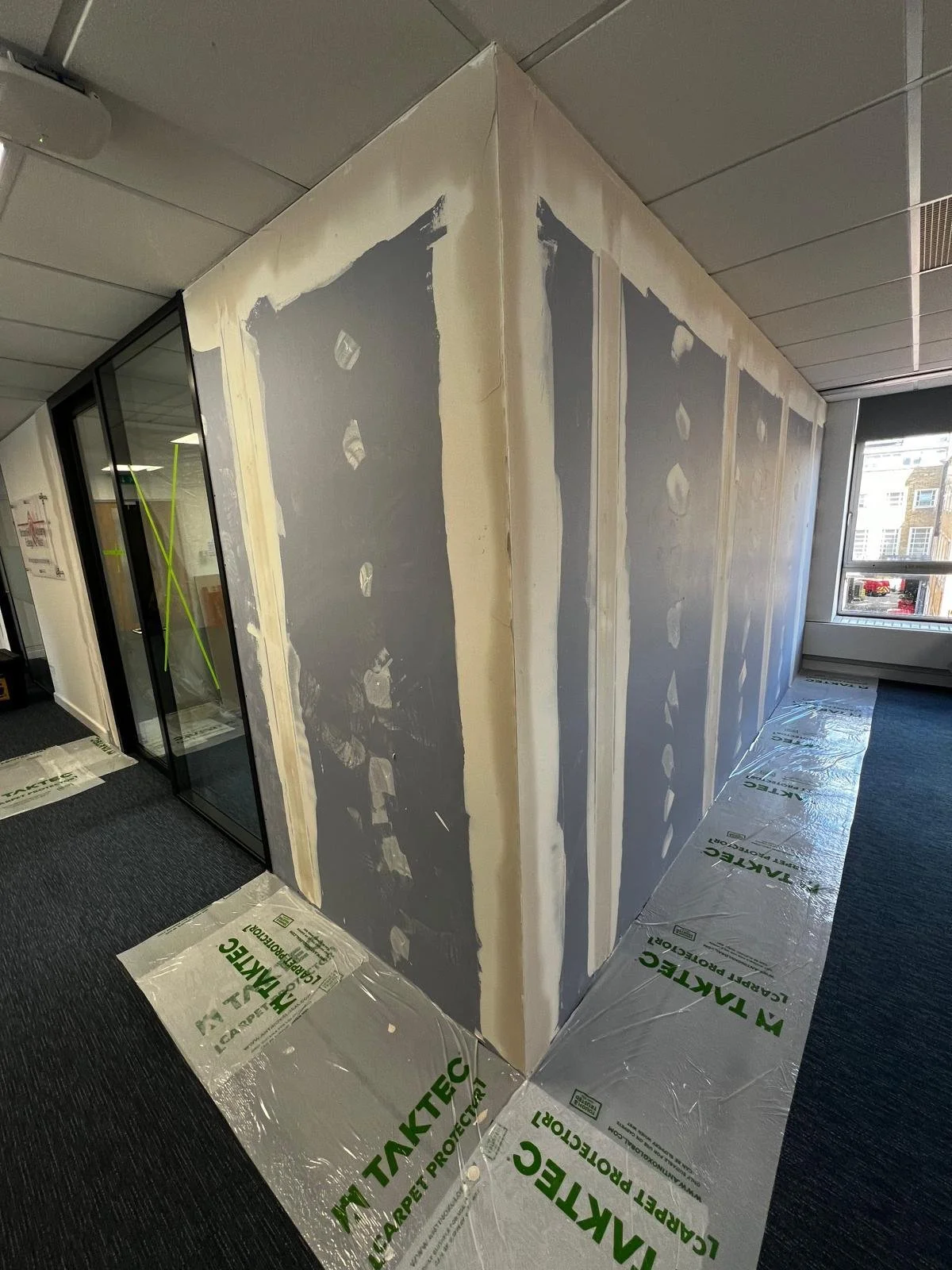 An interior office wall under construction with drywall installed, taped, and mudded, covered with protective plastic on the floor, near a window.