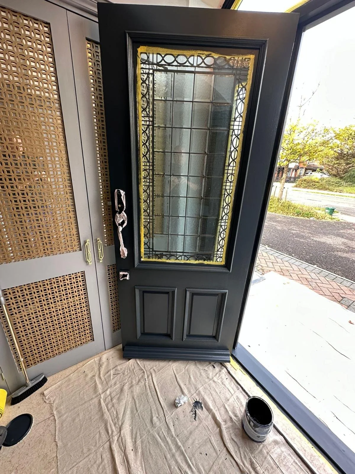 A black front door with decorative glass and trim, partially painted, standing open inside a house. The door is next to a set of beige cabinets with lattice panels and gold handles, with painting supplies and a drop cloth on the floor.