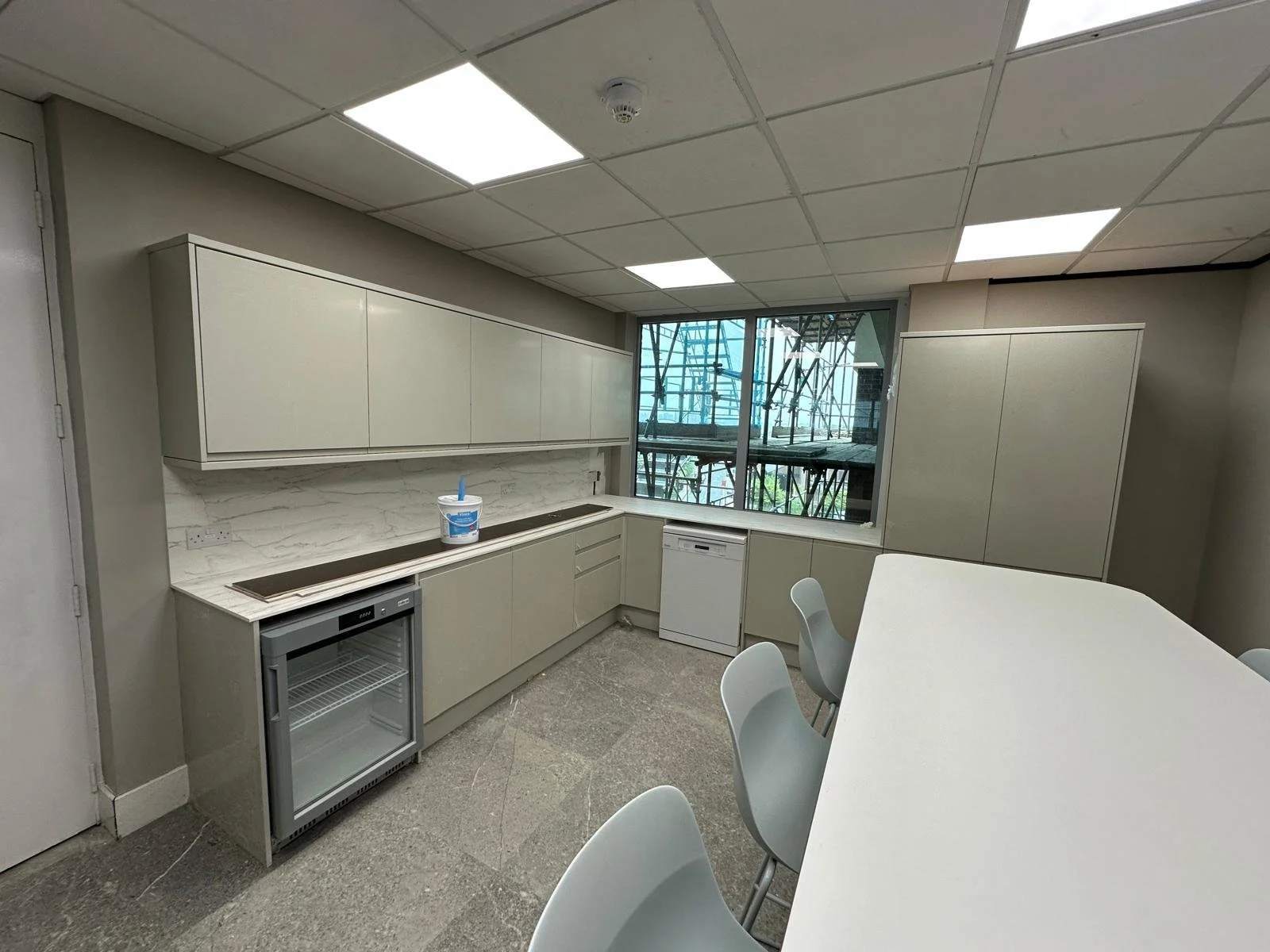 Modern kitchenette with white cabinets, marble backsplash, a small fridge, and a bucket on the counter, overlooking a construction site outside the window.