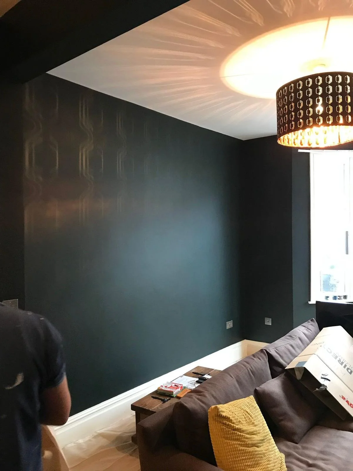 A living room with dark teal walls and a black ceiling, featuring a modern black pendant light and a window with natural light. There is a brown sofa with yellow pillows, a wooden coffee table with miscellaneous items, and part of a person wearing a dark shirt is visible in the foreground.