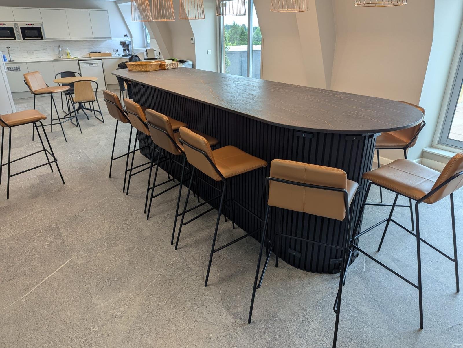 Modern kitchen with a large, curved island with a dark wooden base and a gray marble countertop, surrounded by tan leather barstools. The background features white kitchen cabinets, a coffee machine, and a sliding glass door leading to an outdoor space.