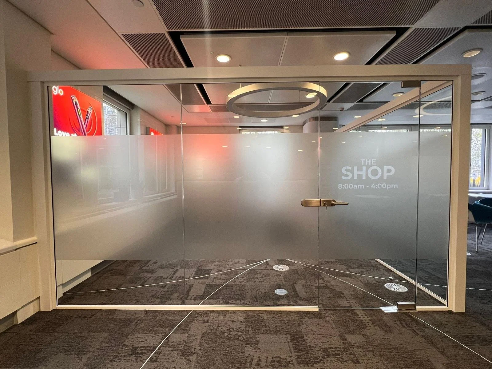 Frosted glass door with text indicating business hours from 8:00am to 4:00pm, in an indoor space with modern ceiling lighting and large windows, possibly an office or retail space.