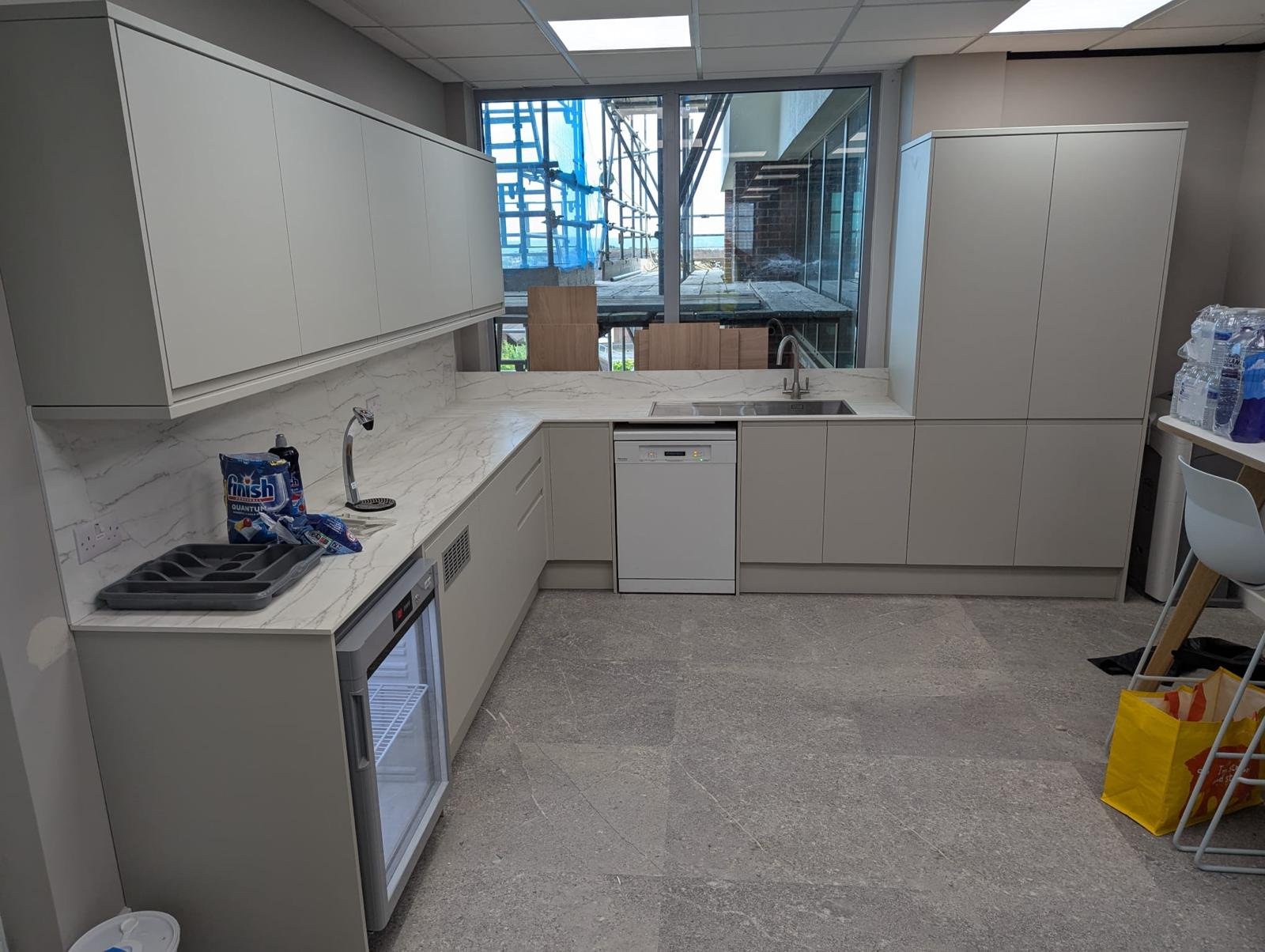 A modern kitchen with white cabinetry, marble countertops, a small sink, a dishwasher, and a mini refrigerator. There are some cleaning supplies and bottled water on a side table, with a window revealing an outdoor construction site.