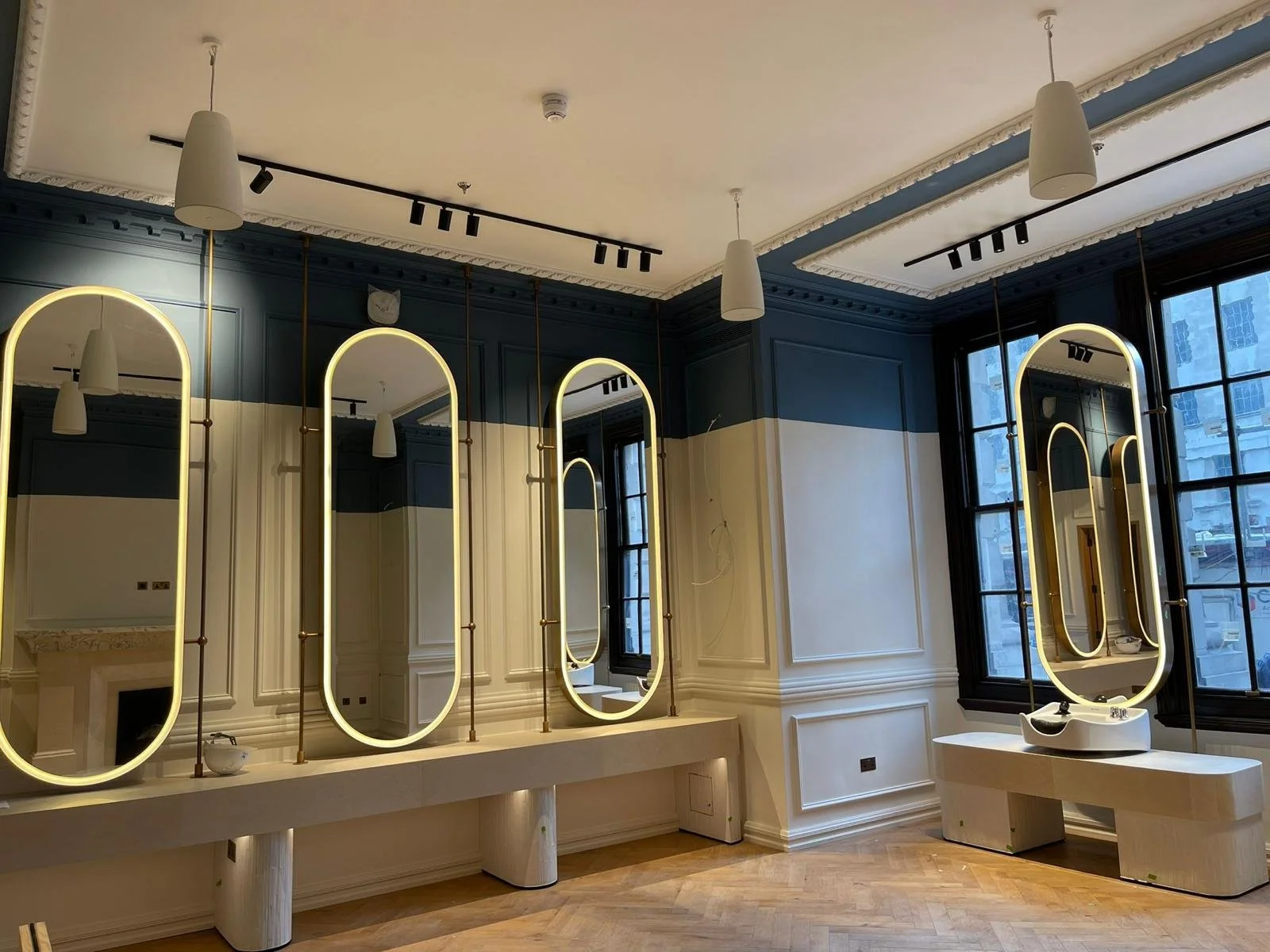 Interior of a modern salon with three illuminated mirror stations, white countertops, and tall black-framed windows with city views. The ceiling features ceiling lights and track lighting, with ornate crown molding.