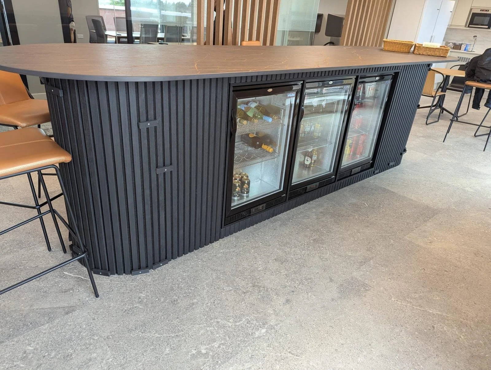 Black beverage cooler with three glass doors filled with drinks, situated on a gray floor in a modern restaurant or café interior.