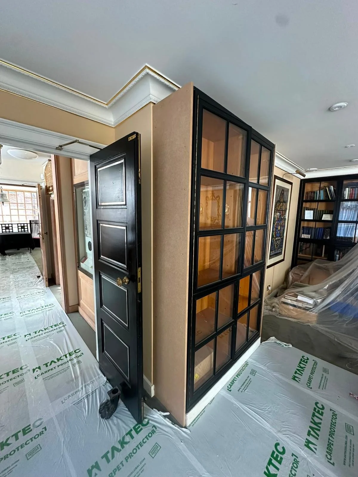 Living room with black-framed glass partition, bookshelves, and partially covered furniture, under renovation with protective floor coverings.