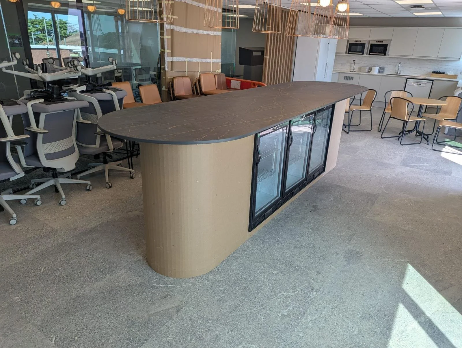 Modern office break room with a large central refrigerator and countertop, surrounded by chairs, a kitchen area with cabinets, microwave, dishwasher, and a sink, and a row of office chairs on the left side.