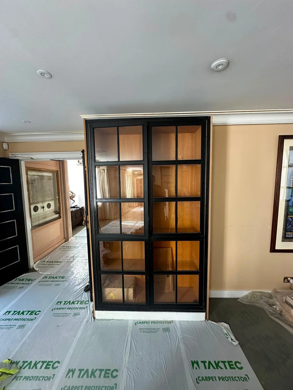 Interior view of a room with a black-framed glass door-shaped bookshelf, wooden shelves inside, beige walls, and a ceiling with recessed lighting. Part of a hallway with a doorway and protective flooring is visible.