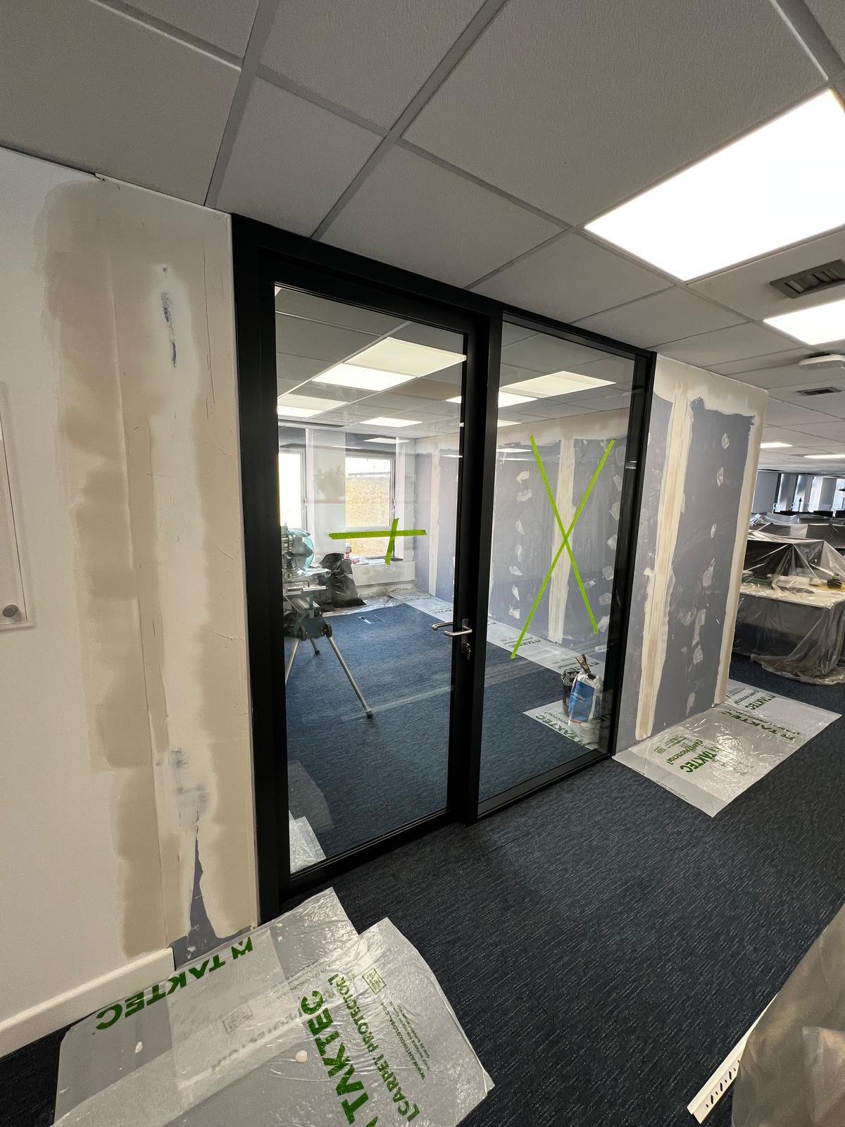 Office space under construction with a glass door, drywall with joint compound, and tools. Green tape and paper on the floor indicate ongoing work.
