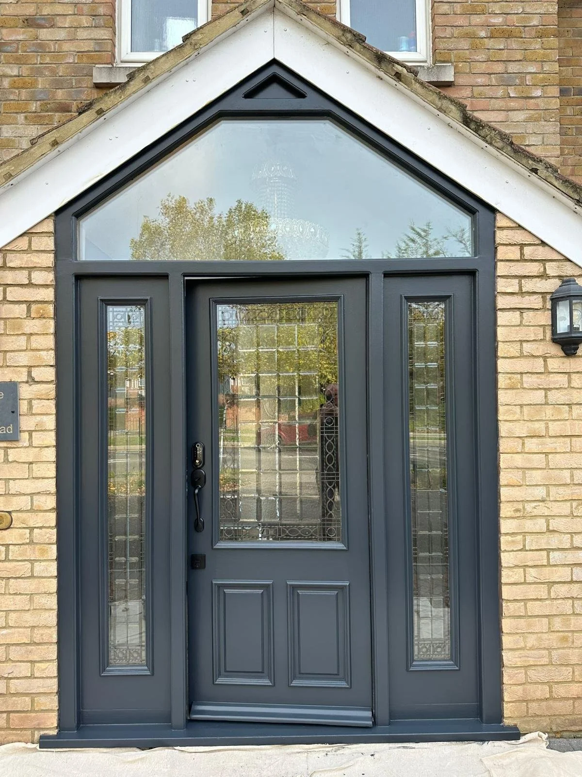 Front door with a dark gray frame and glass panels, set in a brick wall with a large triangular window above.