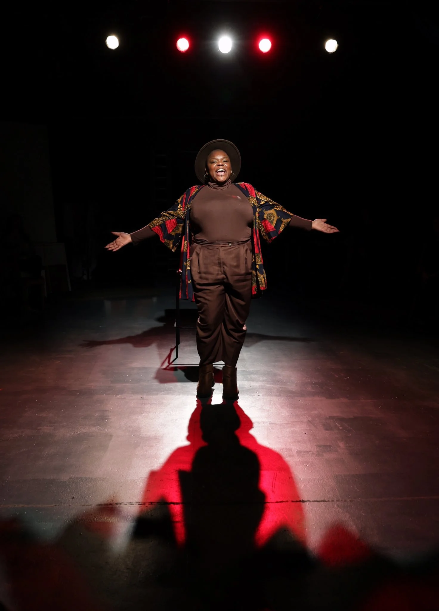 Maz - a fat Black nonbinary person - performing on stage under red and white lights with arms outstretched, casting a shadow on the floor.
