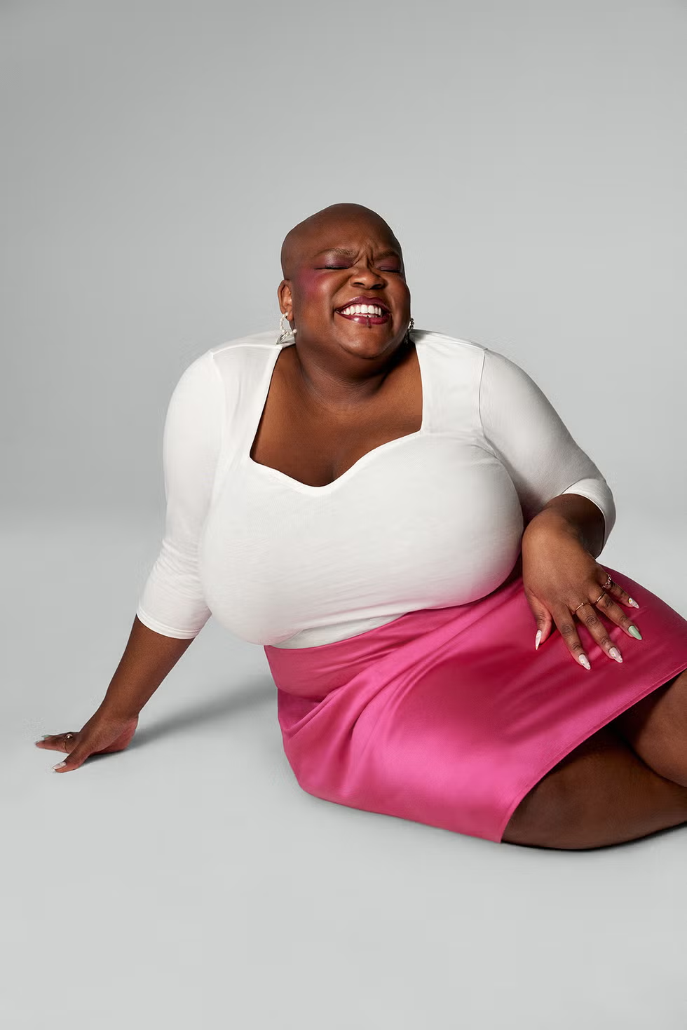 Maz - a fat Black nonbinary person with a shaved head - wearing a white top and pink skirt, sitting on the floor with their legs to the side and one hand resting on the ground.