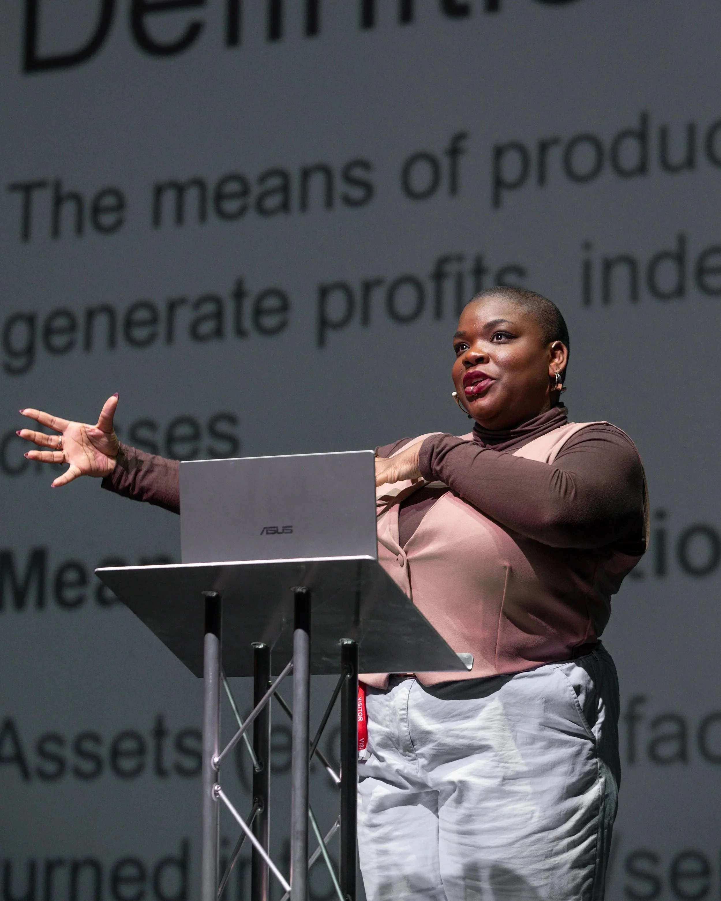 Maz - a fat Black nonbinary person - speaking behind a lectern, gesturing with both hands