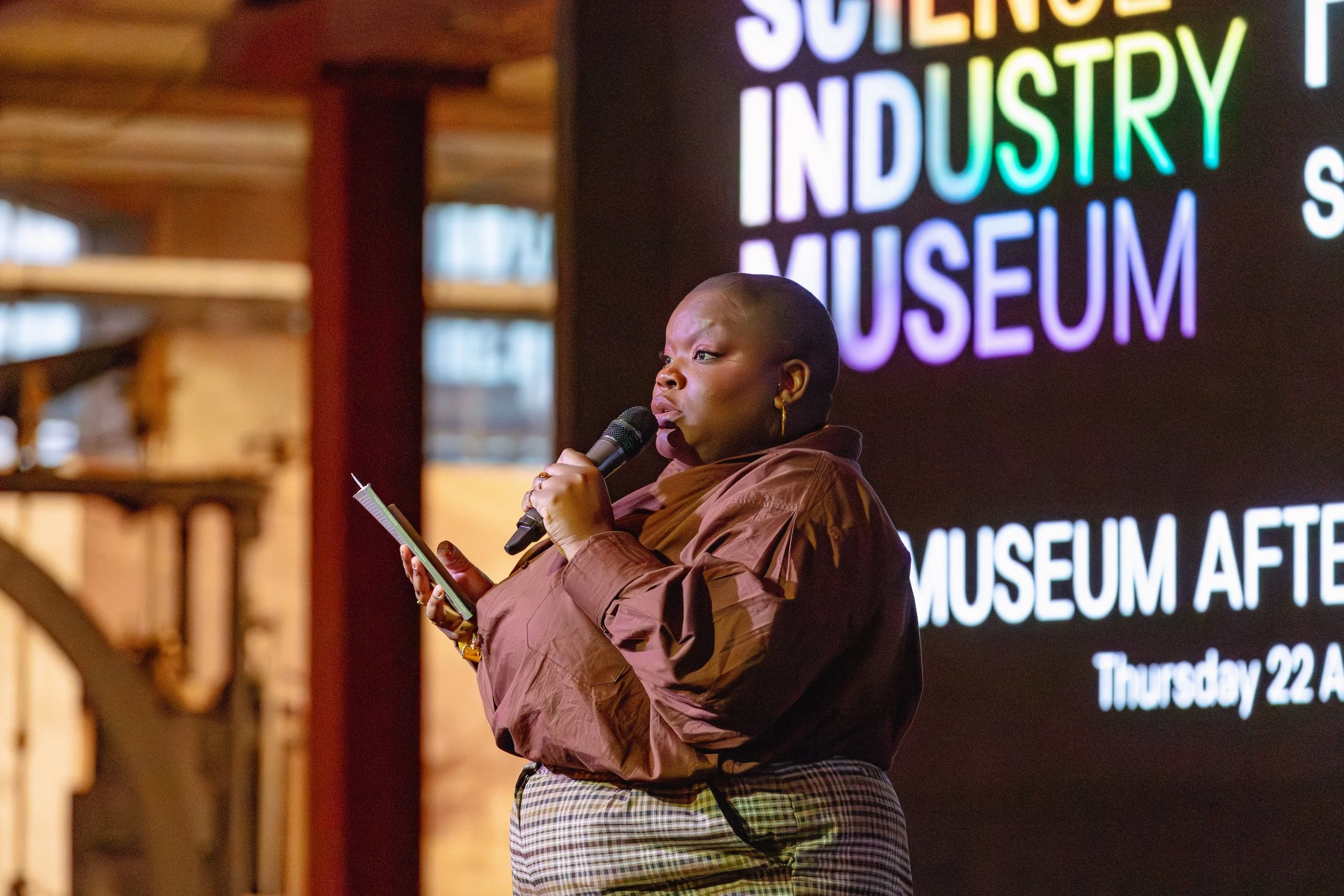 A woman speaking into a microphone during a presentation at an industry museum event, with a large illuminated sign behind her reading 'SOUTHERN INDUSTRY MUSEUM'.